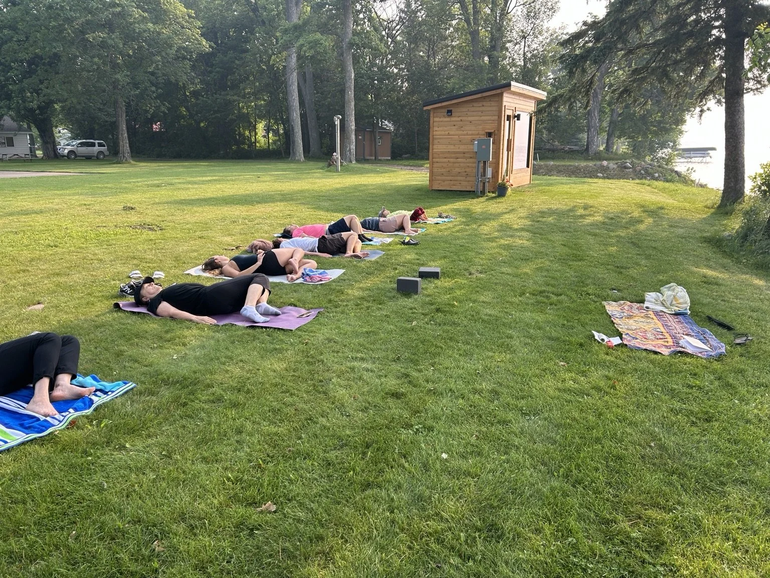 Group of people practicing stillness and breath practices outdoors on a grassy area with mats, near trees and Mille Lacs lake in Minnesota.