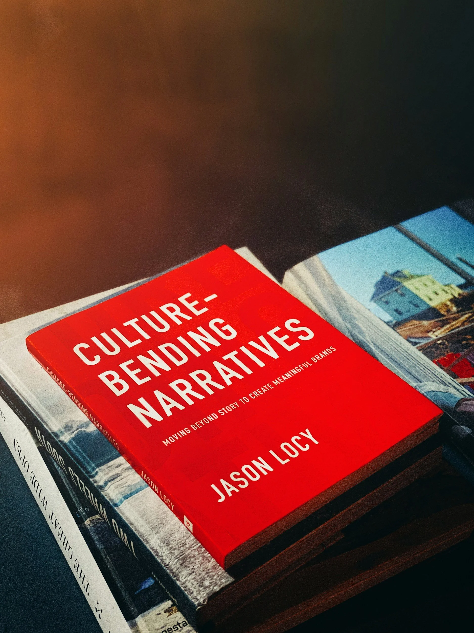 Close-up of a red book titled "Culture-Bending Narratives" by Jason Loxy on a coffee table, with a part of another magazine visible underneath and a cityscape photograph in the background.
