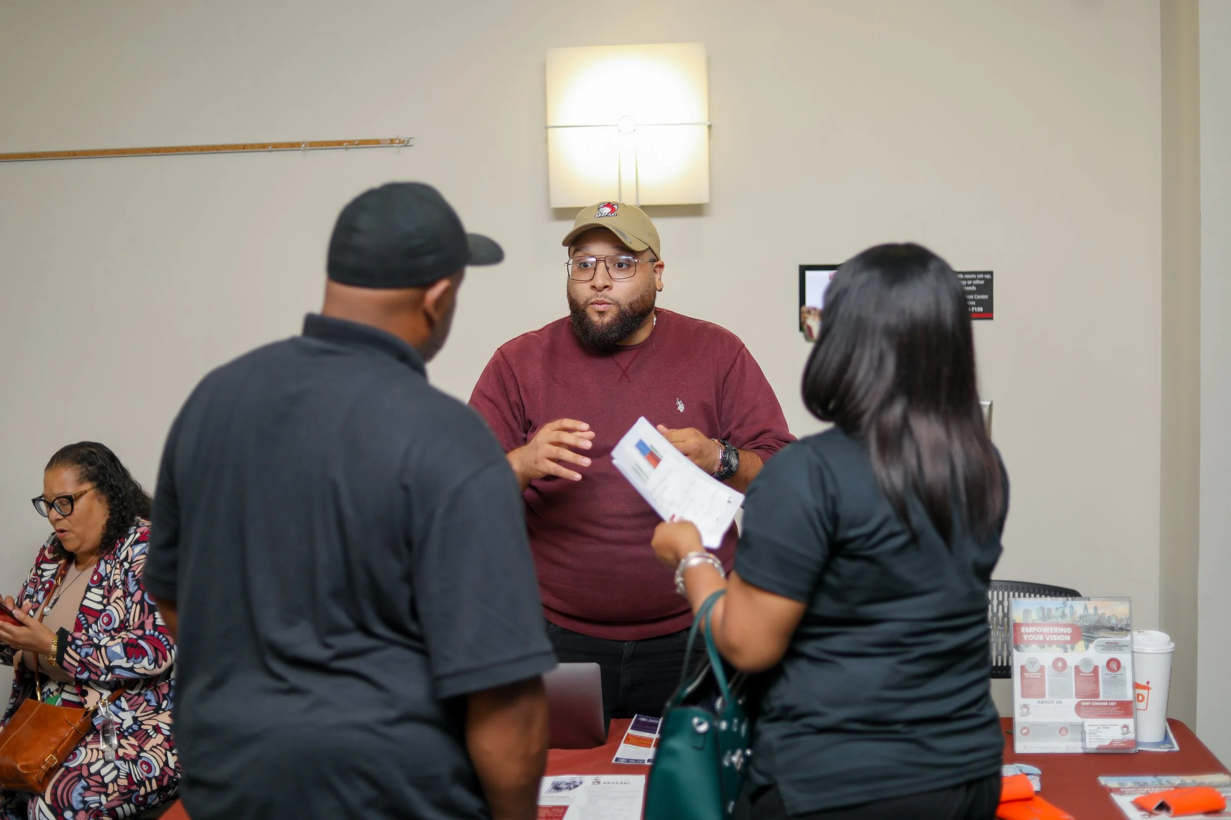 Three people engaged in conversation at a booth, with a woman sitting and using her phone in the background.