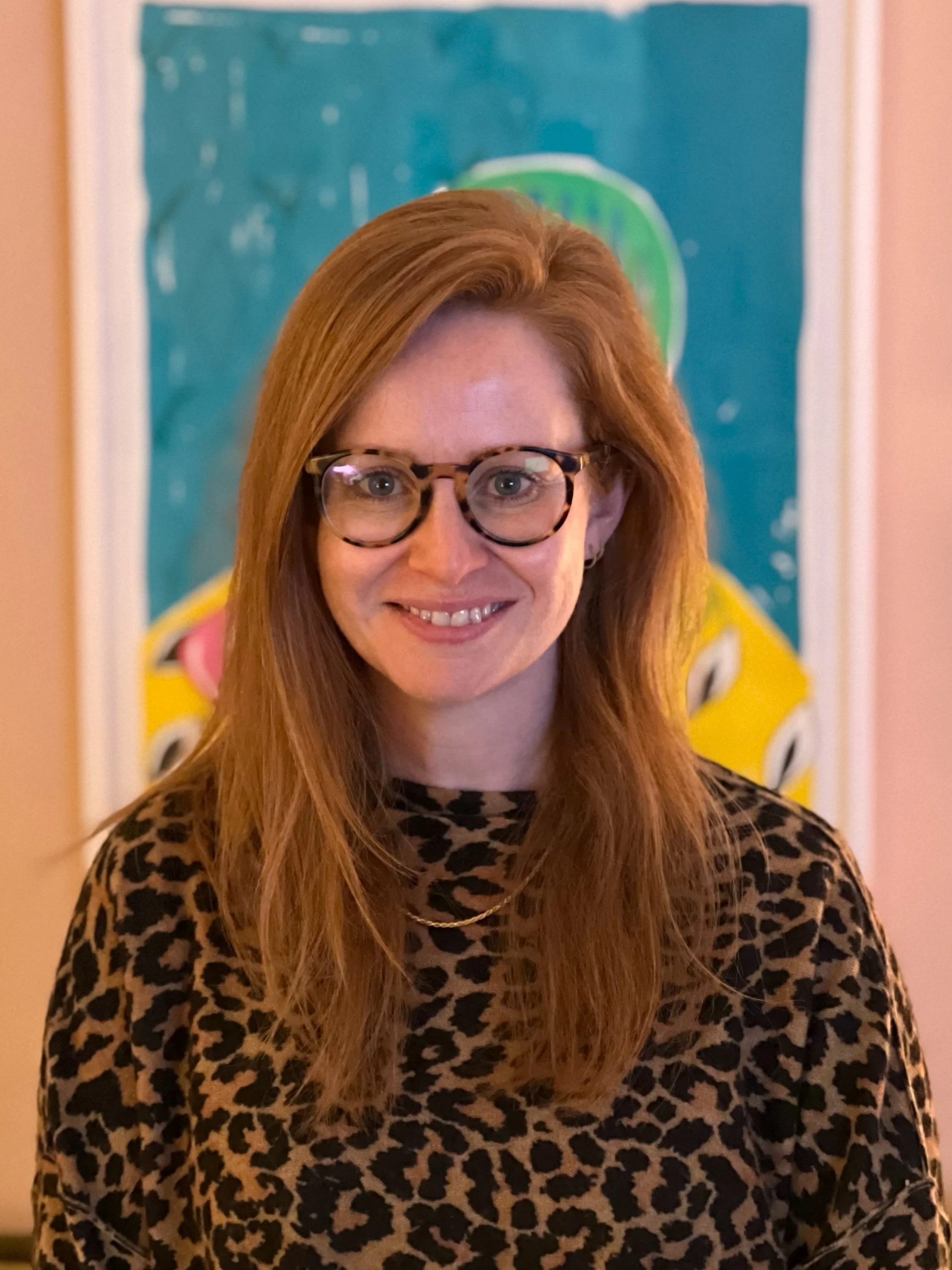 A woman with long red hair and glasses, smiling, wearing a leopard print top and standing in front of colourful artwork.