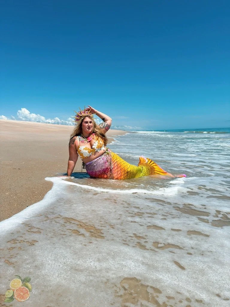 A person dressed as a mermaid with a yellow and pink tail and floral top, wearing a crown, sitting on a sandy beach in the shallow water with their arm raised, under a clear blue sky.