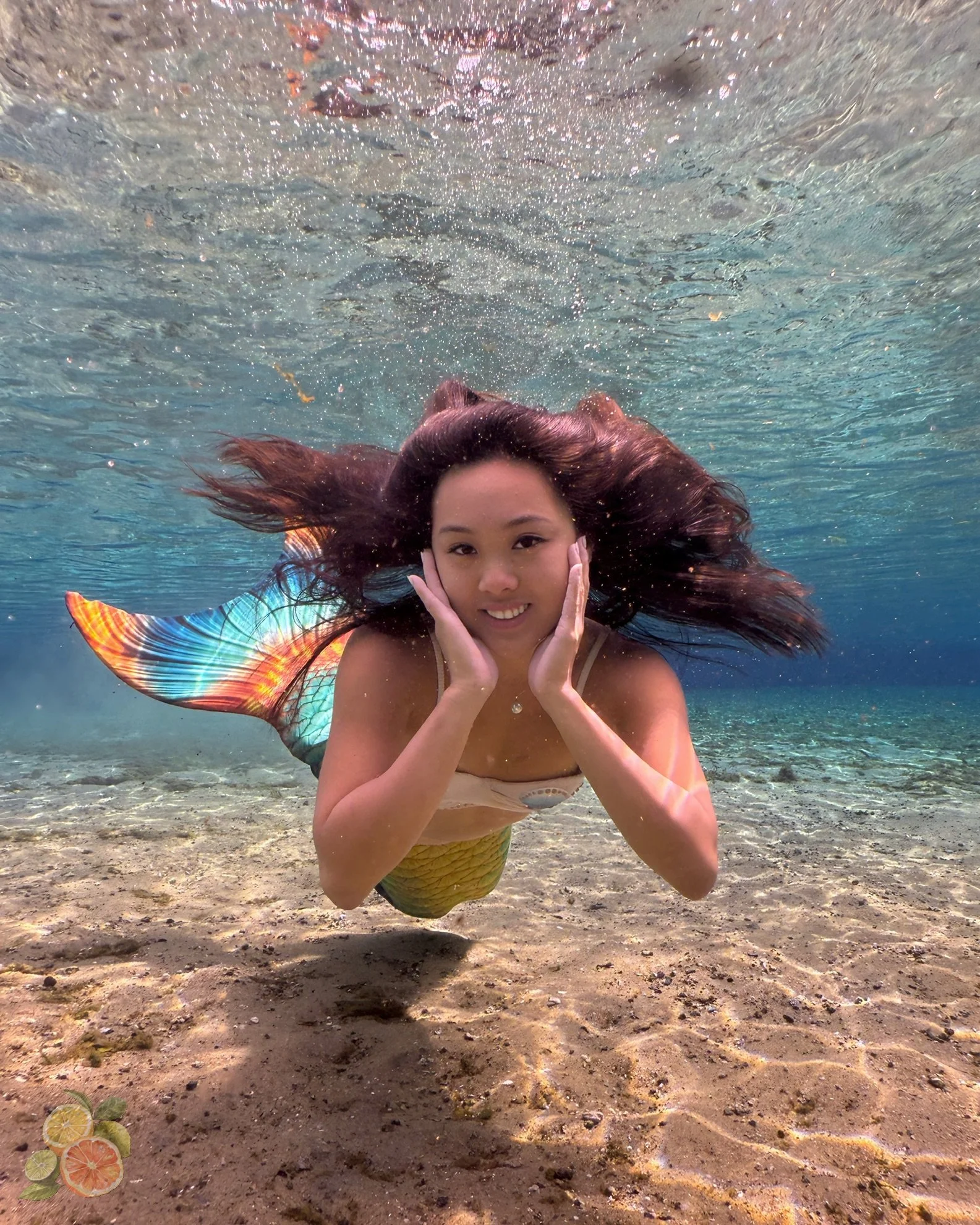 A woman with long dark hair in a mermaid tail swimming underwater and holding her face with both hands.