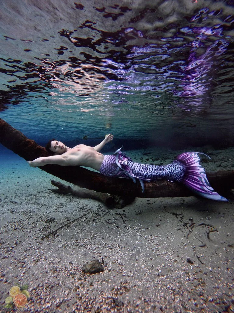 A person with a mermaid tail lying on a submerged log in an underwater scene, with water rippling above and sandy bottom beneath.