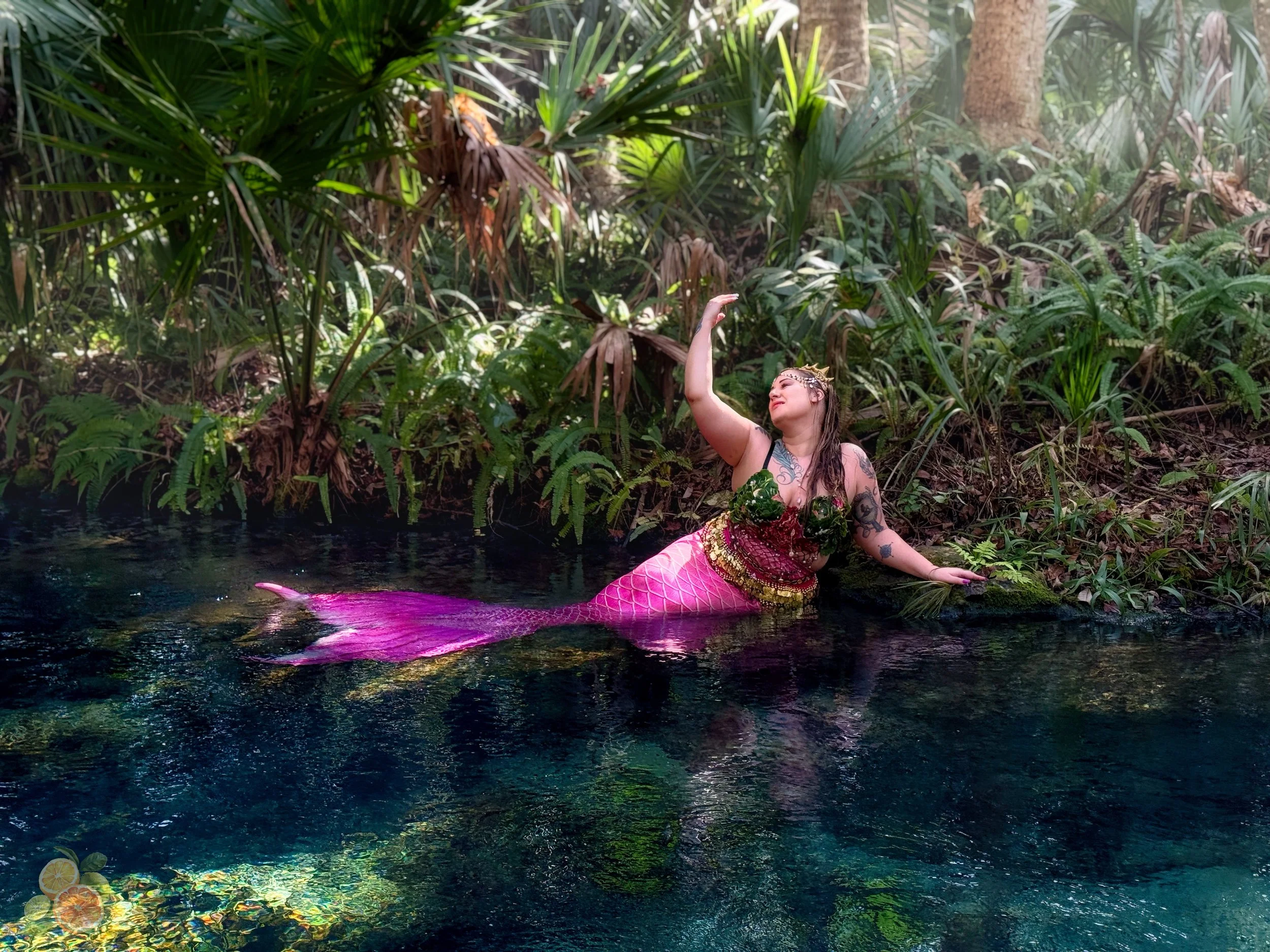 A woman dressed as a mermaid sitting in a forested riverbank, wearing a sequined green and pink top with a pink scale-patterned tail, surrounded by lush greenery and water.