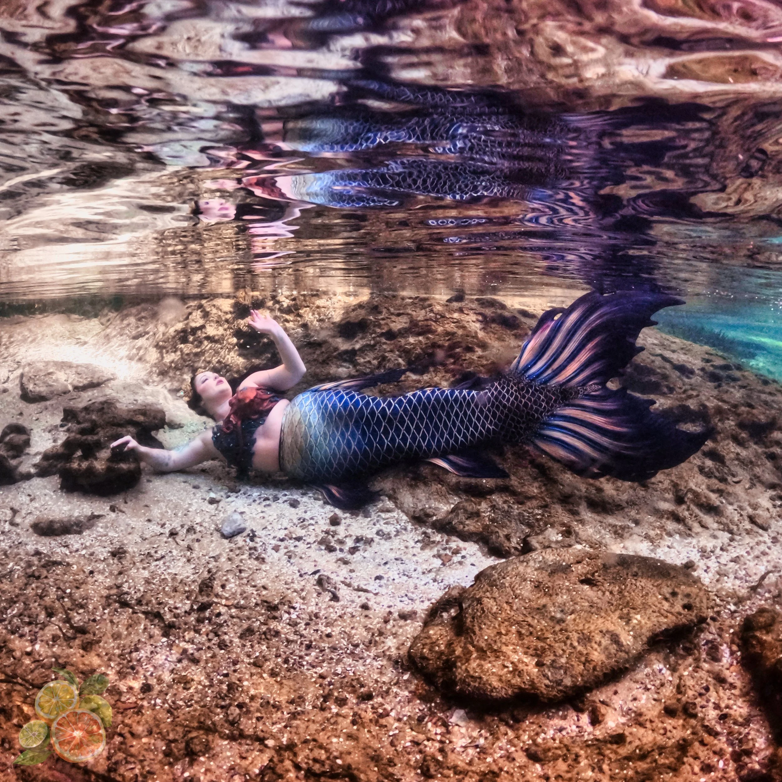 A woman dressed as a mermaid with a patterned tail and top, lying on a rocky surface underwater, with rocks and water surrounding her.