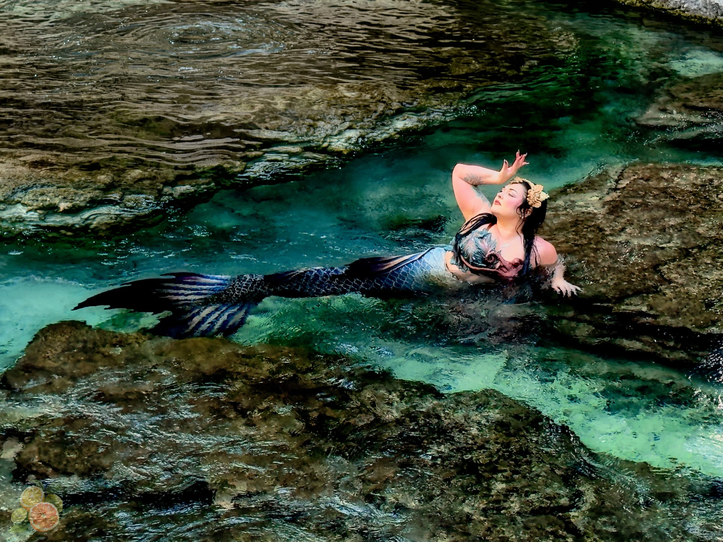 A woman with tattoos and a floral headband dressed as a mermaid, lying on rocks in a small pool of clear turquoise water, raising one hand above her head with eyes closed.