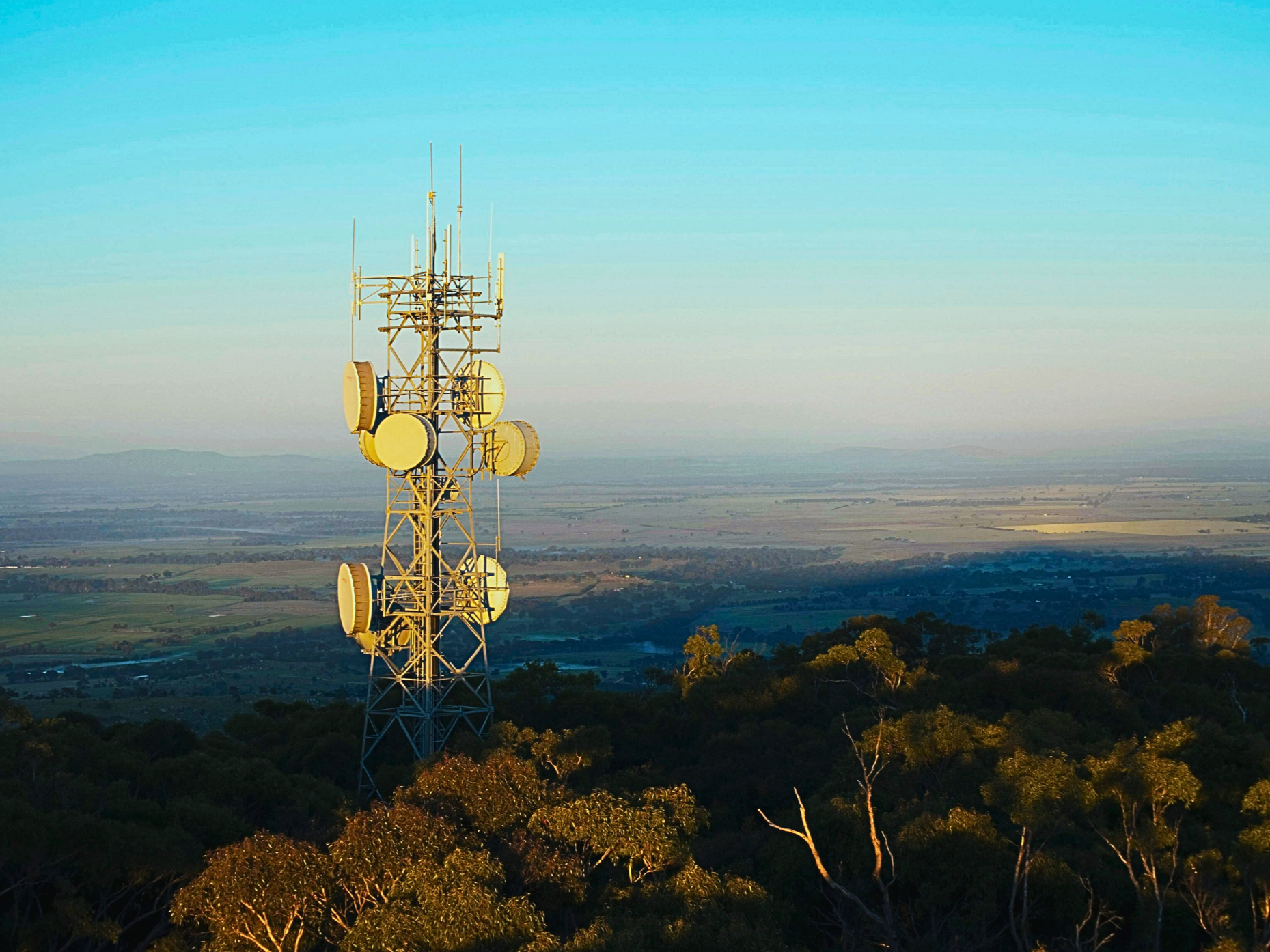 A tall telecommunications tower with multiple satellite dishes and antennas against a partly cloudy blue sky.