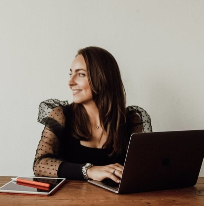 A woman with long brown hair sitting at a wooden table with a laptop, a notebook, and a pen, smiling and looking to her left.