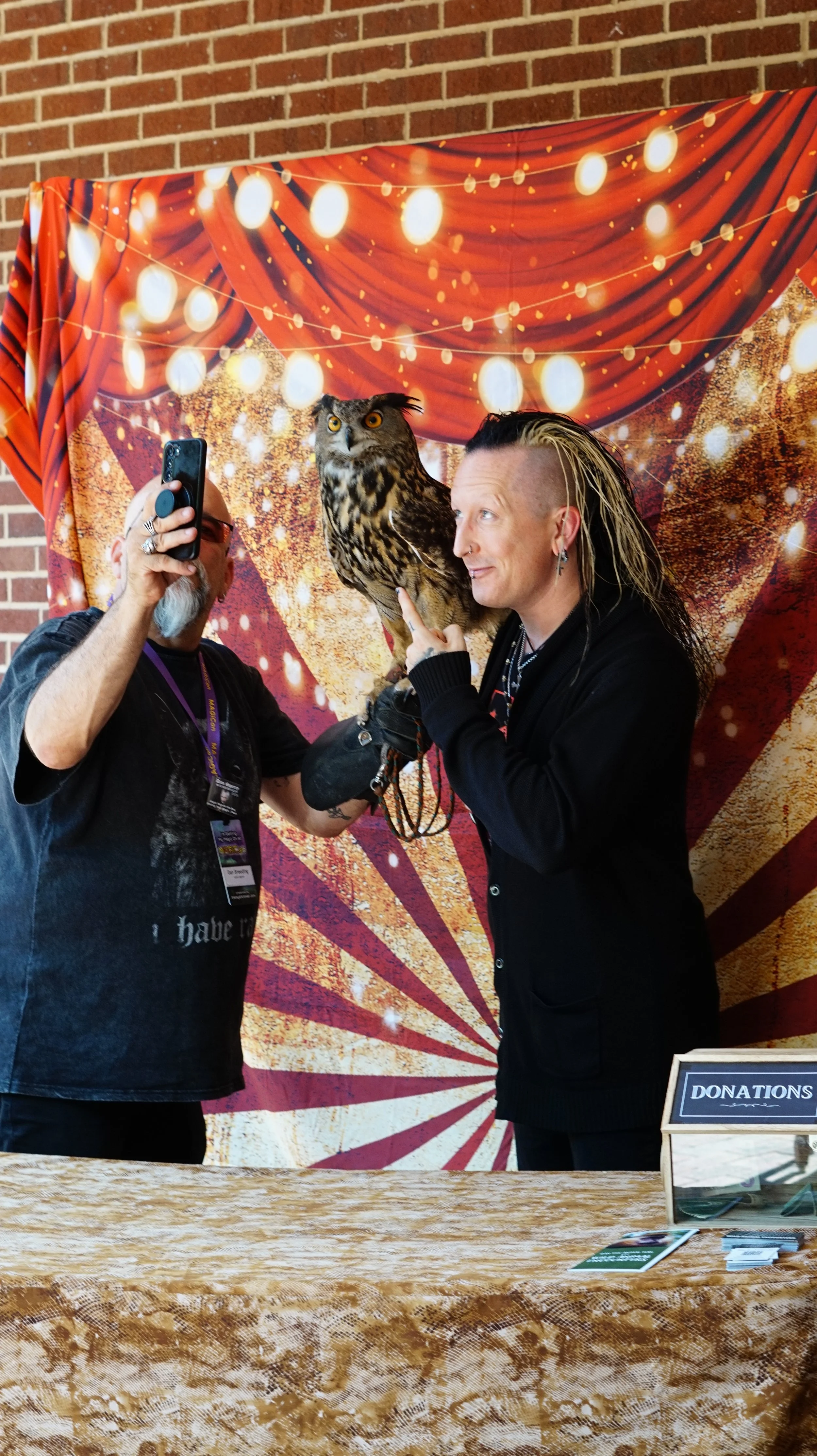 A person with a mohawk hairstyle holding an owl on their hand, and another person taking a photo with a phone, in front of a festive backdrop with red drapes and string lights.