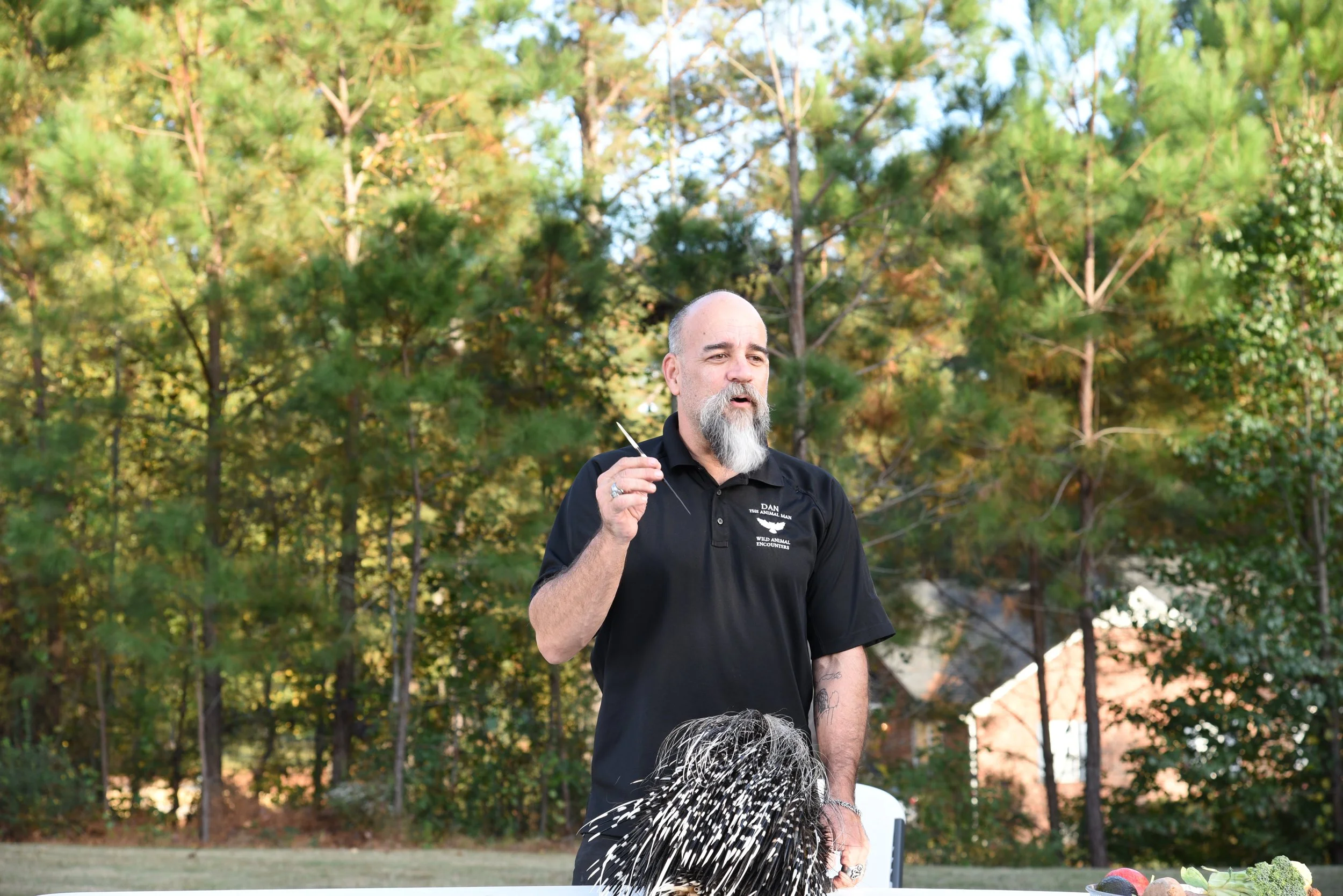 A man with a gray beard and balding head standing outdoors among trees, holding a string in his right hand, wearing a black shirt with text