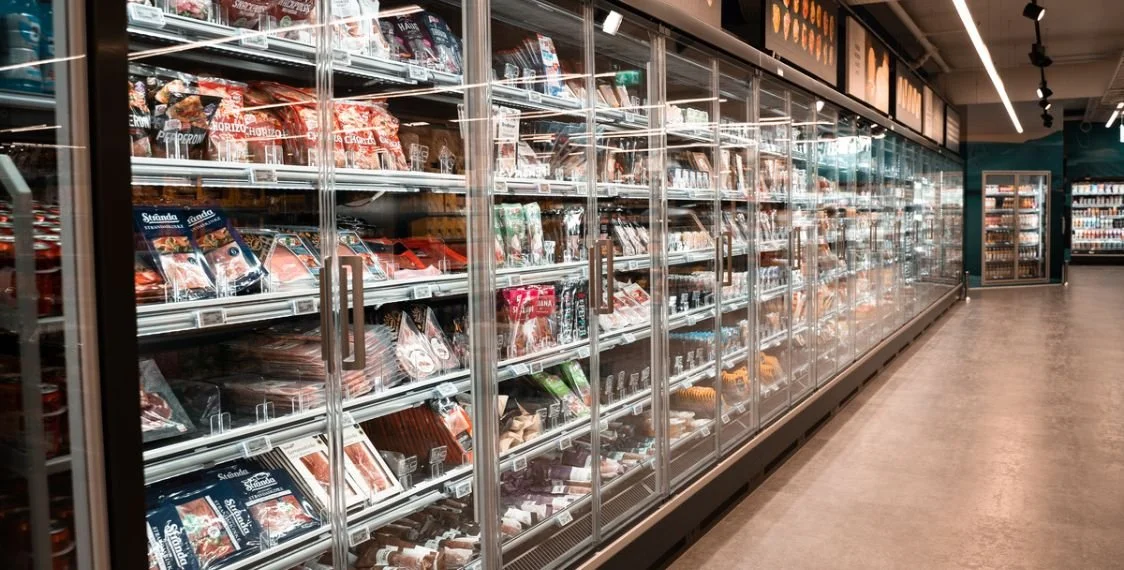 Frozen food aisle with glass-door freezers filled with various packaged meats and frozen products in a grocery store.