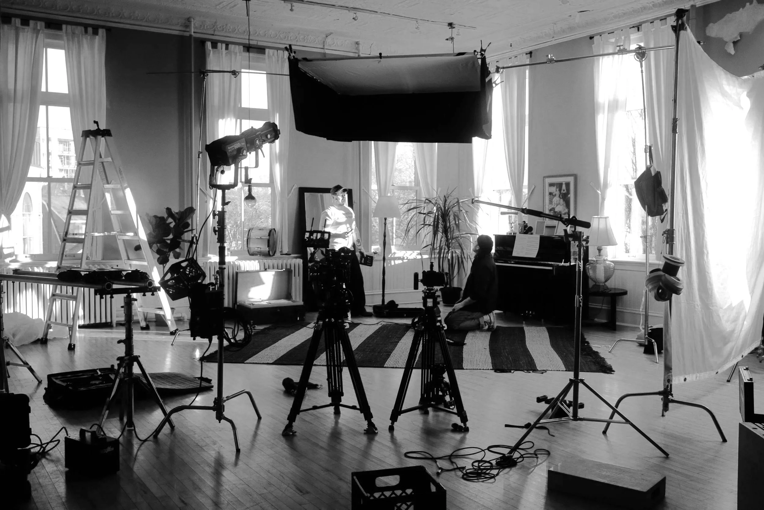 Indoor film or video shoot setup with two people, cameras, lighting equipment, ladder, curtains, and a piano in a room with large windows.