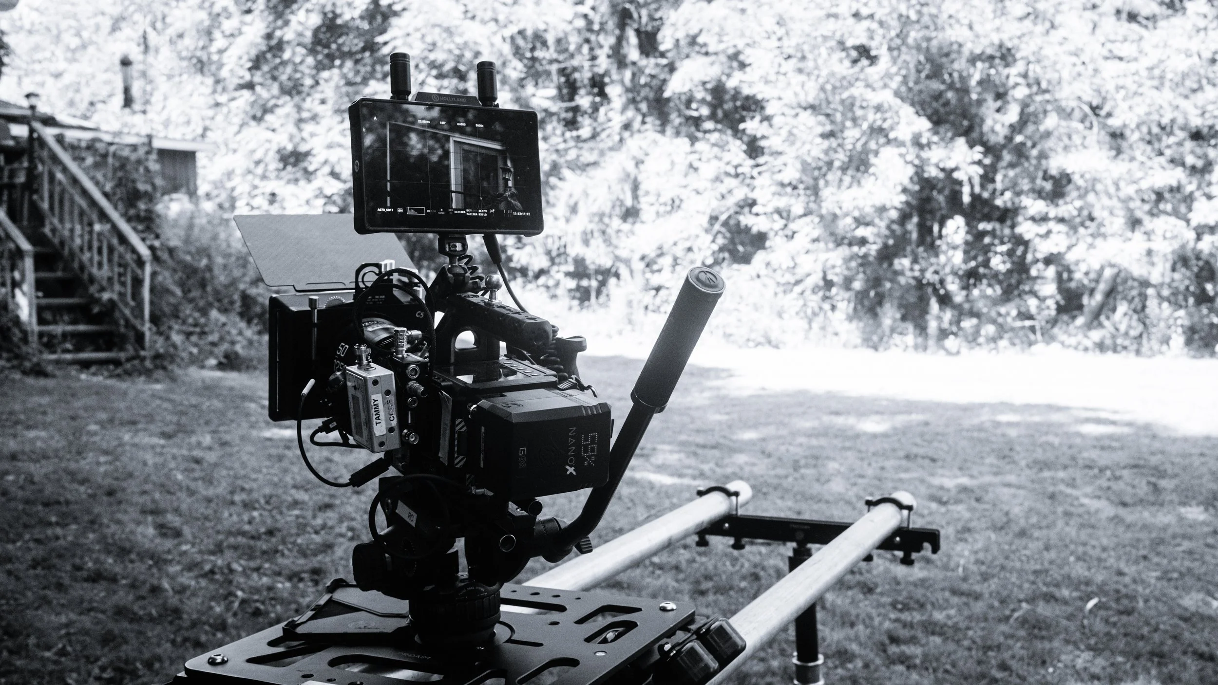 A professional video camera mounted on a rig outdoors in front of trees and grass, with a monitor displaying the camera view.