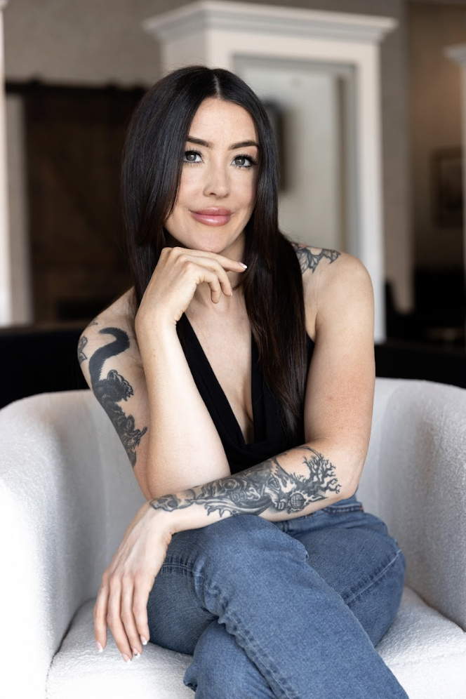 A woman with long dark hair and tattoos on her arms, dressed in a black top, sitting on a black chair against a plain light-colored background.