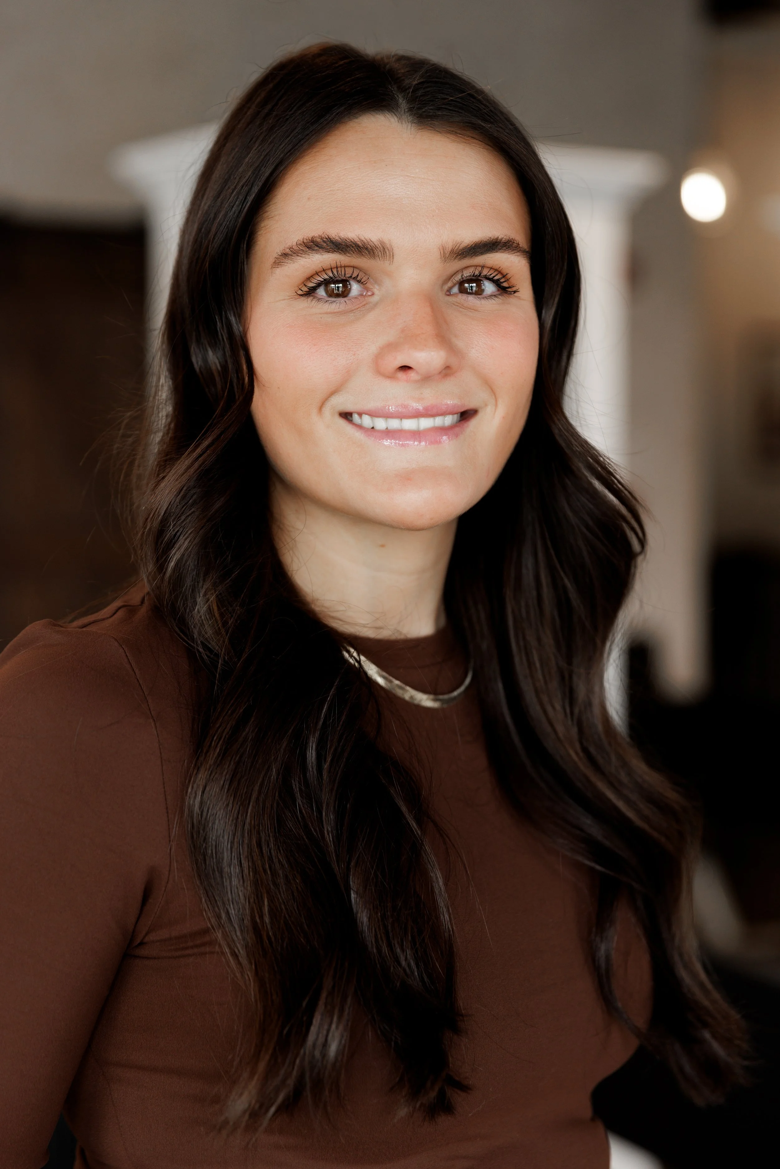 A woman with long wavy brown hair, wearing a black shirt and black ripped jeans, sitting on a beige tufted sofa inside a furniture store.