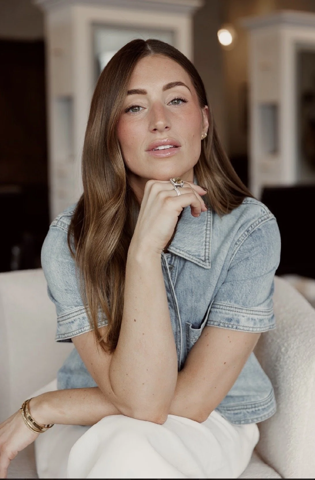 A woman with long wavy light brown hair smiling, wearing a beige button-up dress with puffed sleeves, standing against a plain neutral background.