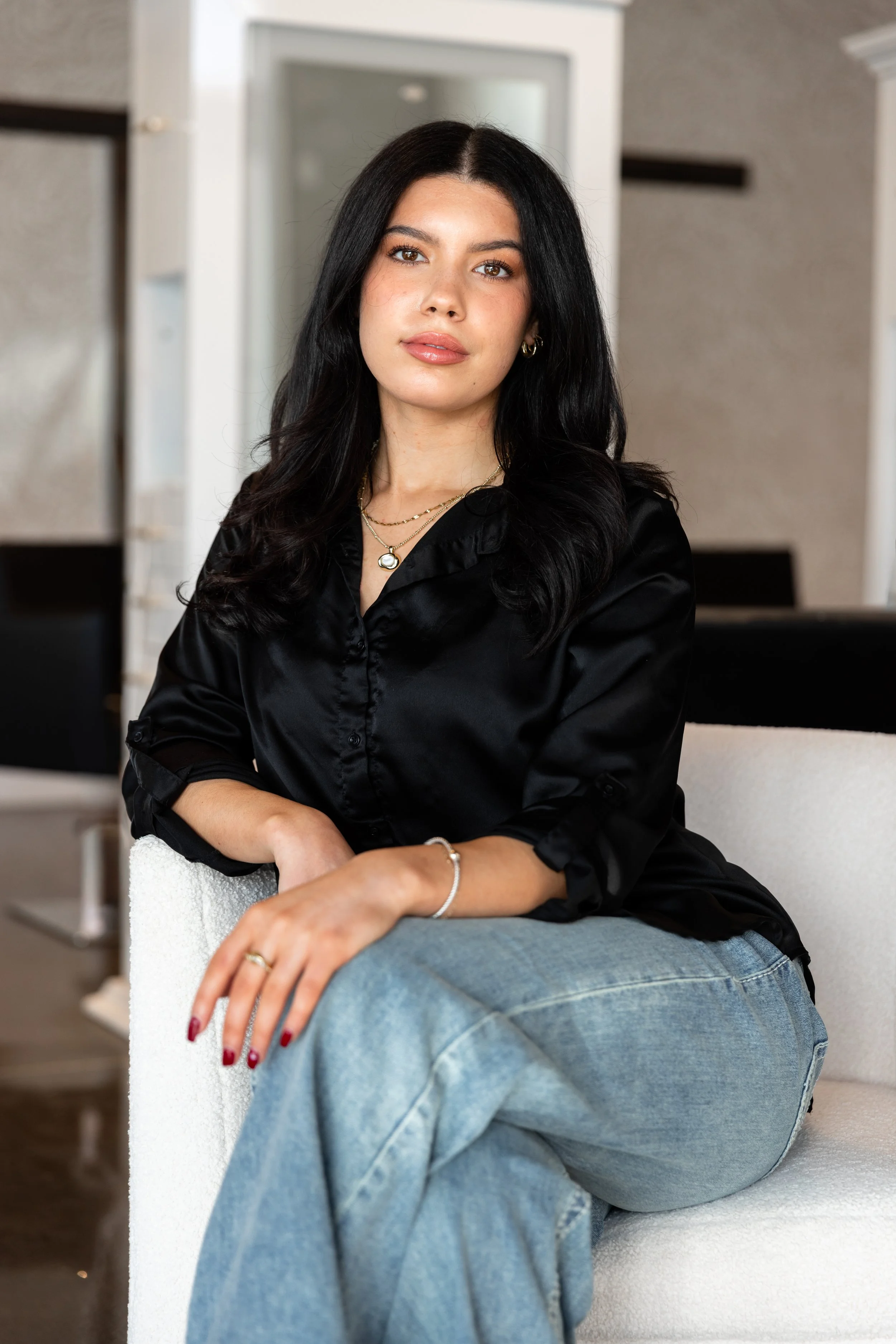 A young woman with medium-length wavy brown hair, wearing a black sleeveless top and light blue high-waisted jeans, standing indoors in front of a patterned beige wallpaper and a wooden door.