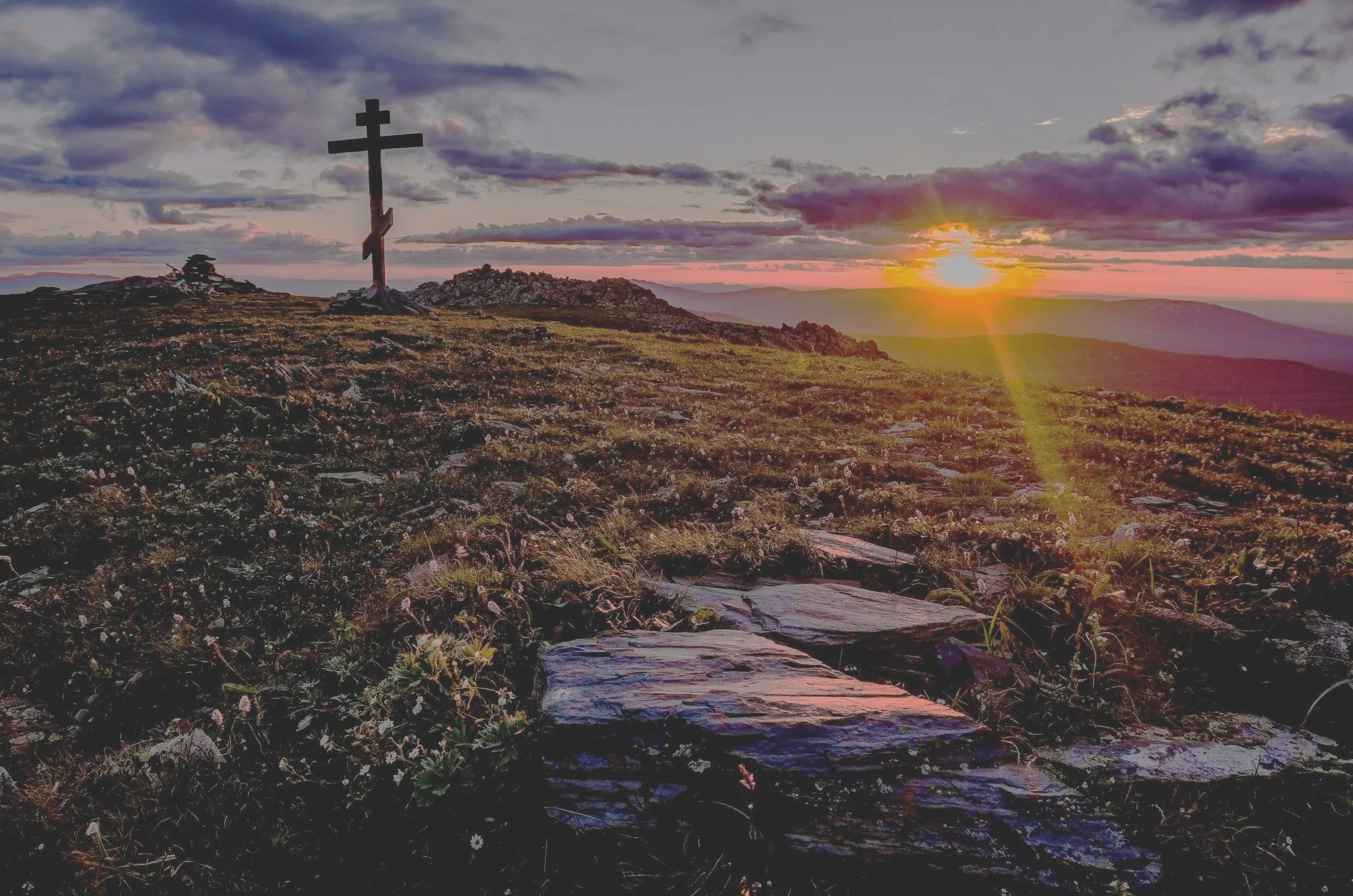 A sunset over a mountain landscape with a large crucifix at the top of a hill, a rocky path leading toward it, and clouds in the sky.