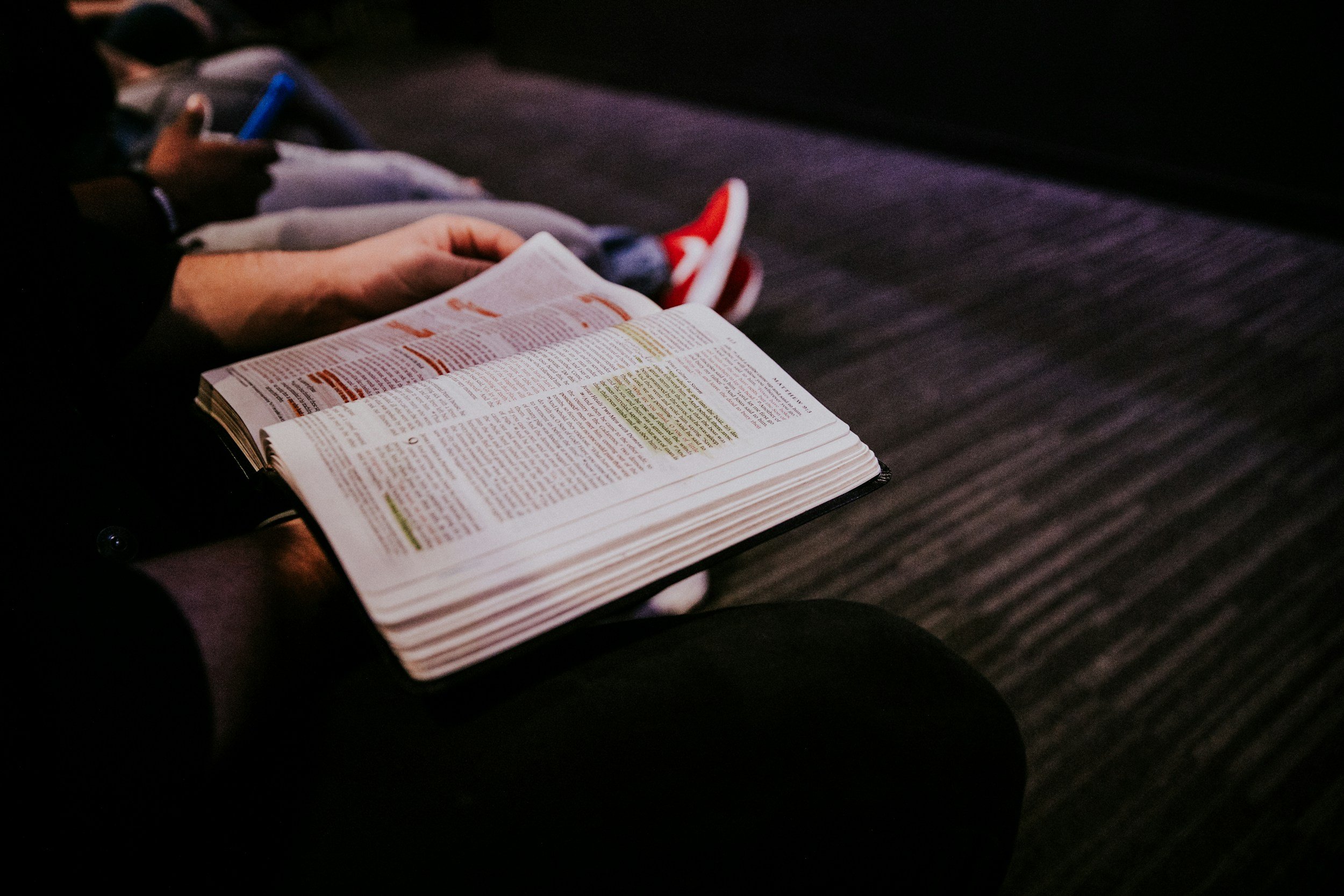 A person holding an open Bible with highlighted passages, seated on a dark room or auditorium floor, with other people sitting nearby.