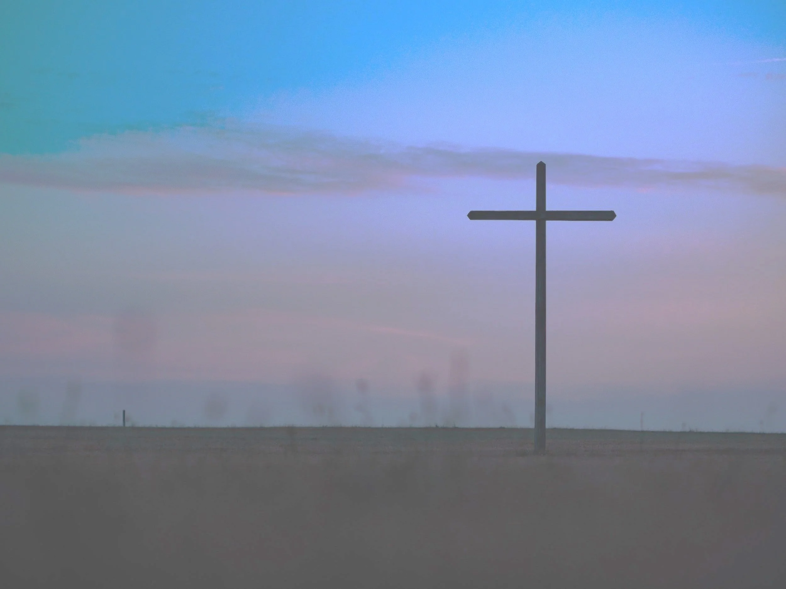 A solitary cross standing in an open field during dusk, with a sky filled with pastel-colored clouds.