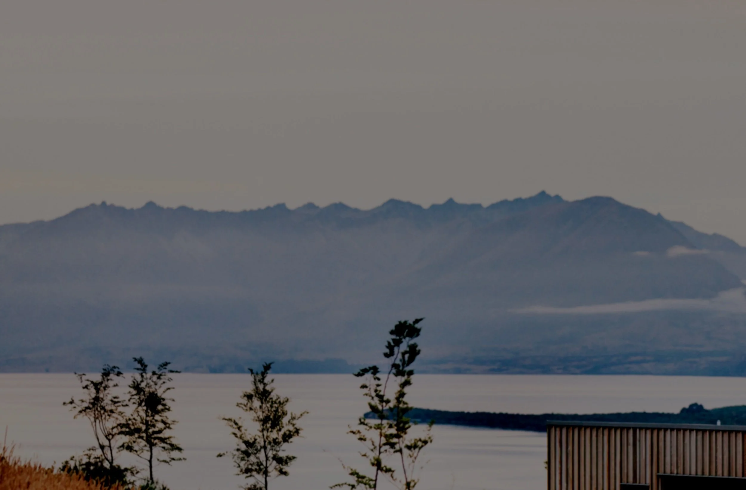 A landscape with mountains in the background, a body of water, a few trees in the foreground, and a wooden structure to the right.