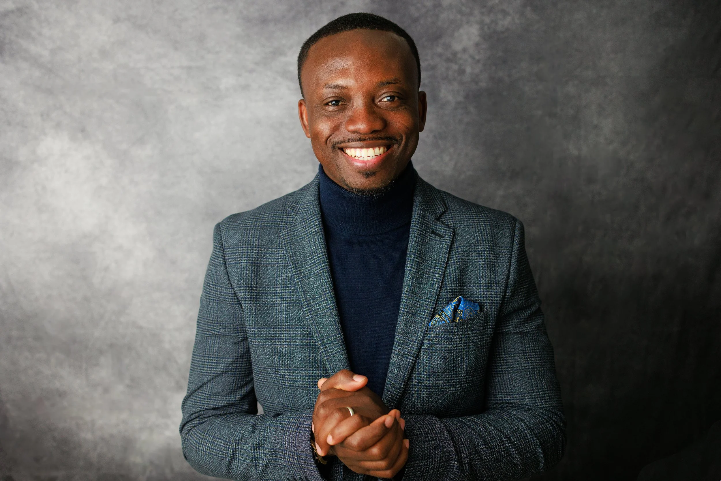 A smiling man in a dark turtleneck and plaid blazer with a blue pocket square, standing against a gray background.