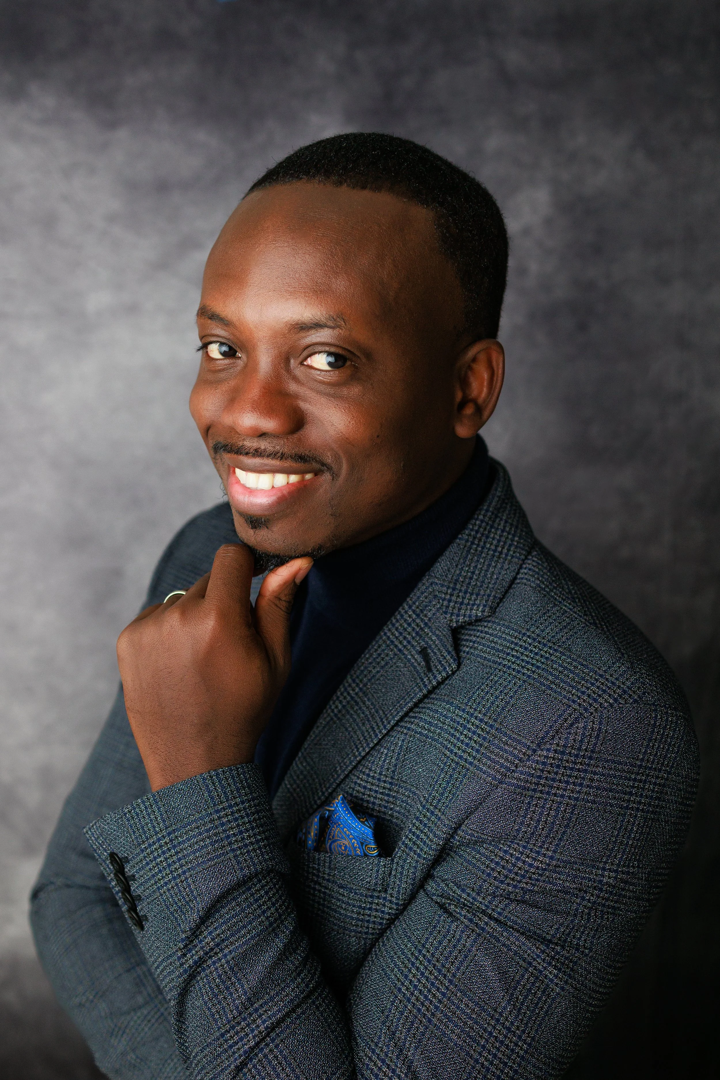 Professional man in a plaid blazer smiling, with one hand on his chin, against a gray background.
