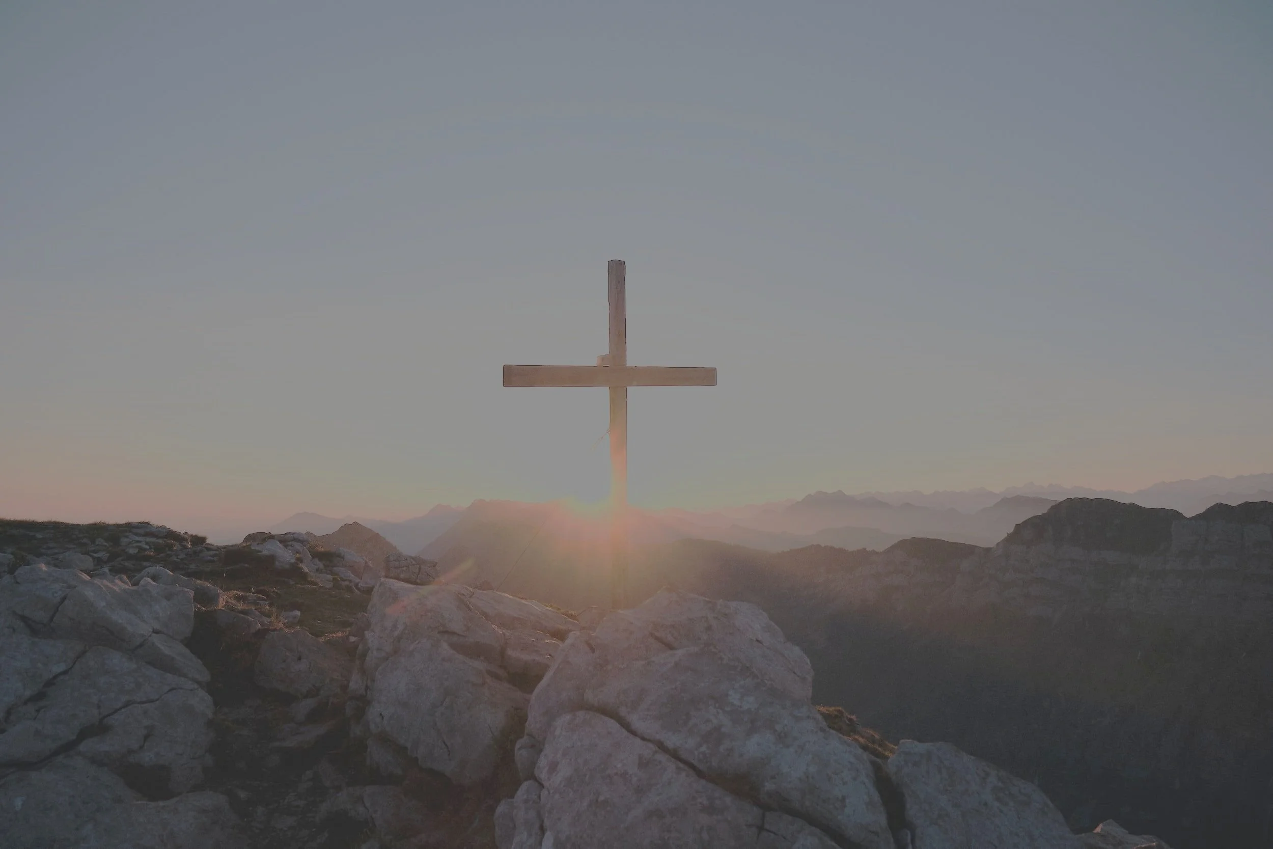 A wooden cross on a mountain summit with a sunrise or sunset, rocky terrain in the foreground, and mountain ranges in the distance.