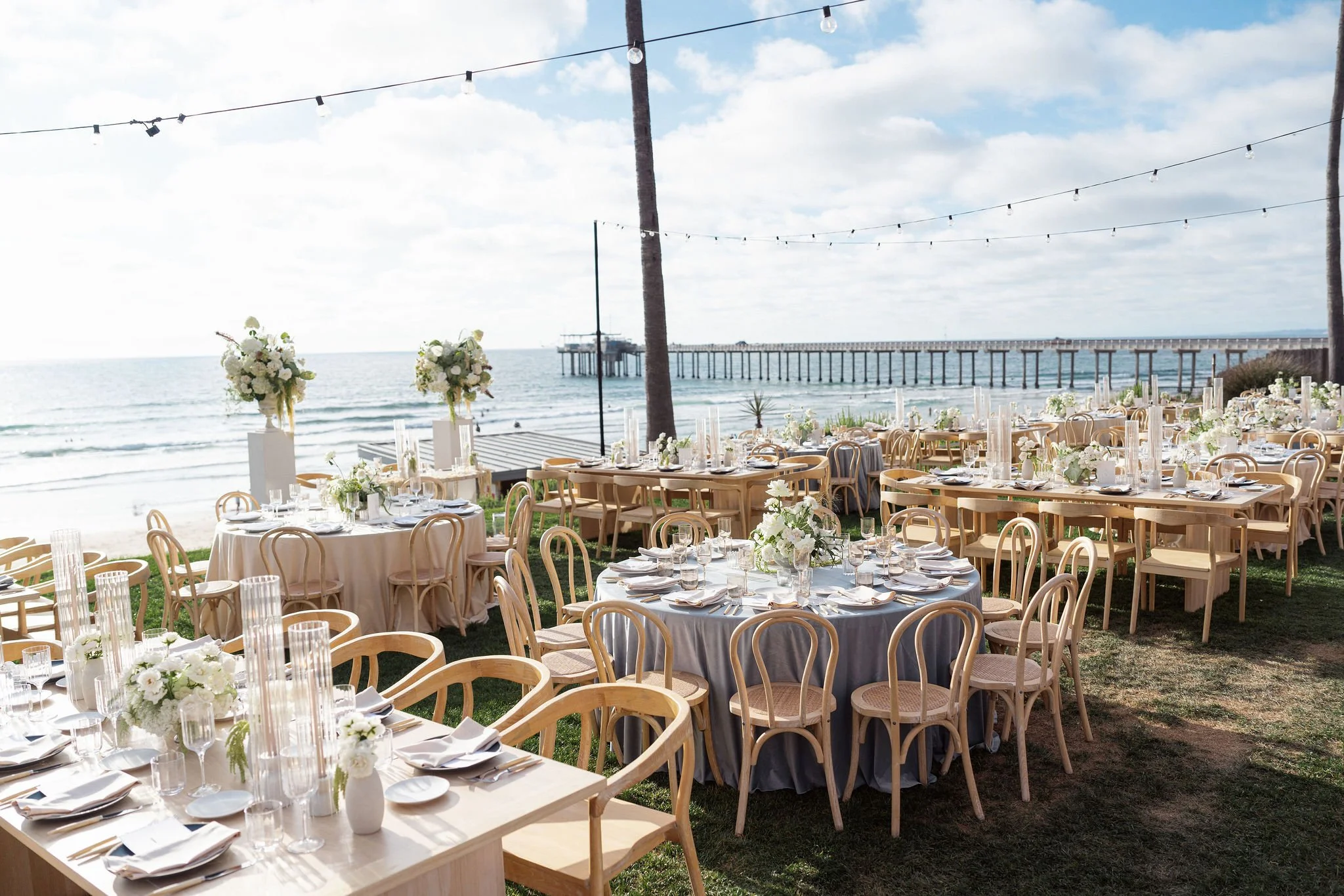 Photo of a beachfront wedding venue set for a wedding reception that is coastal inspired. It has a view of the La Jolla pier and has a mix of long and round tables.
