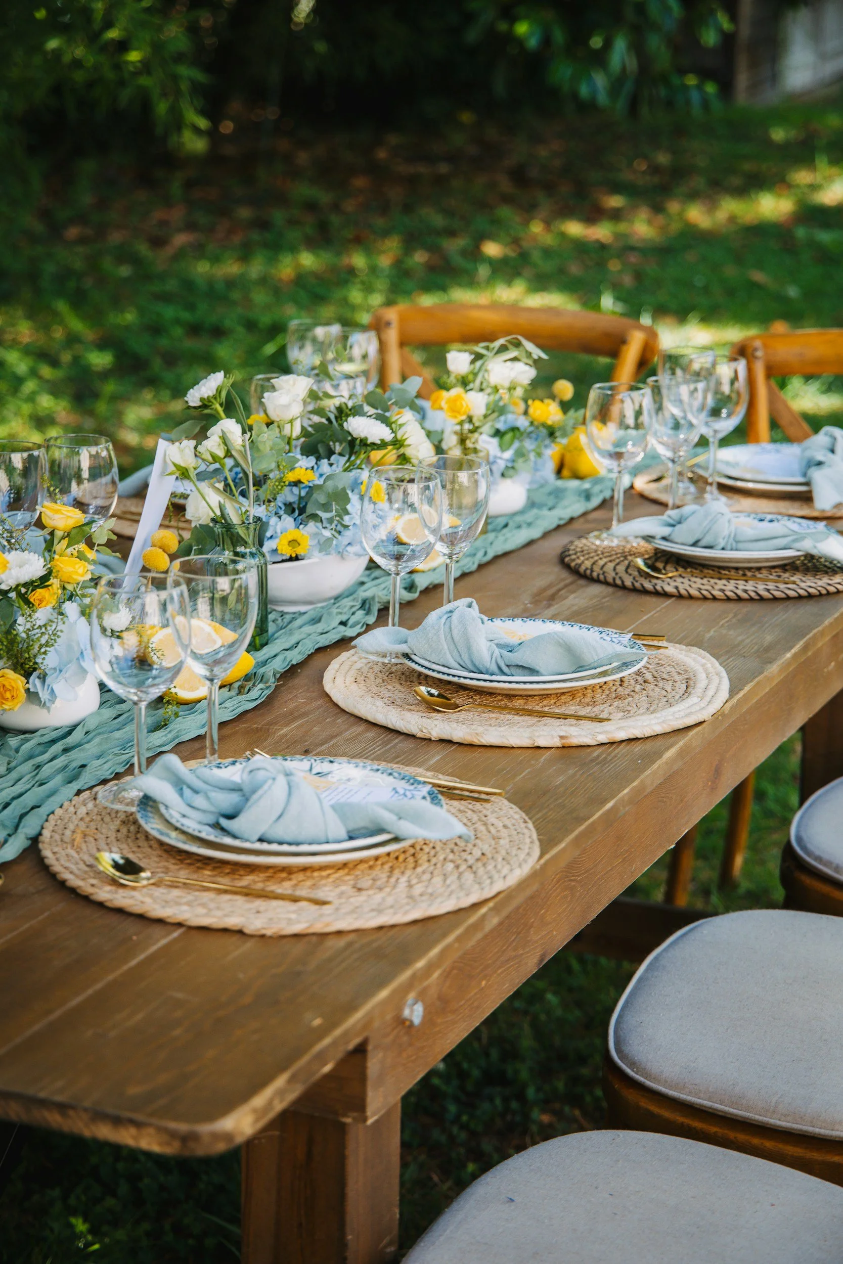 An outdoor dinner table setup featuring a light blue runner, woven rattan placemats, elegant glassware, and fresh summer floral arrangements with yellow and white blossoms.