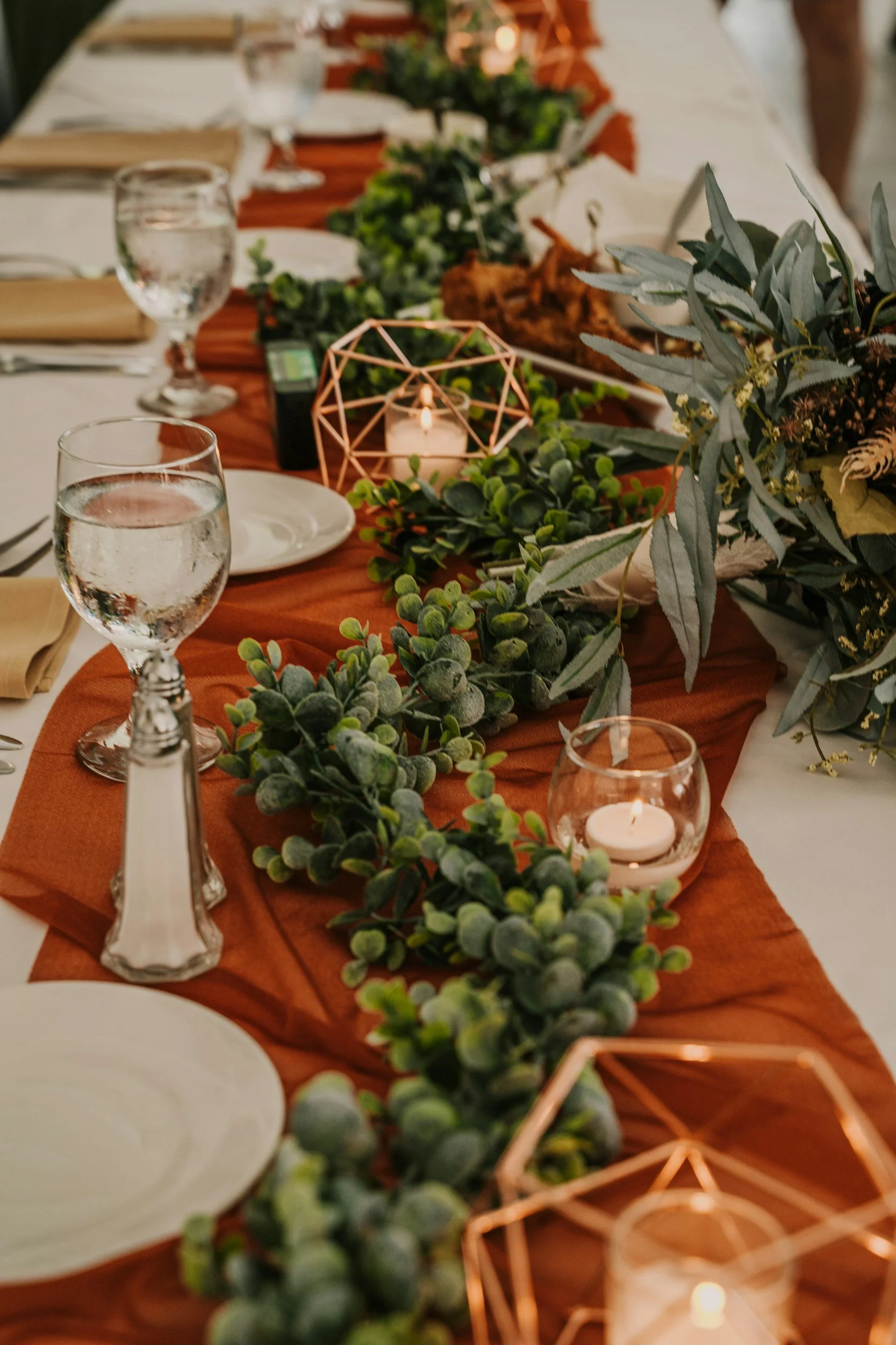 An autumn dinner table setting with a warm terracotta runner, glowing candlelight, leafy green garlands, and wine glasses for an intimate evening gathering.