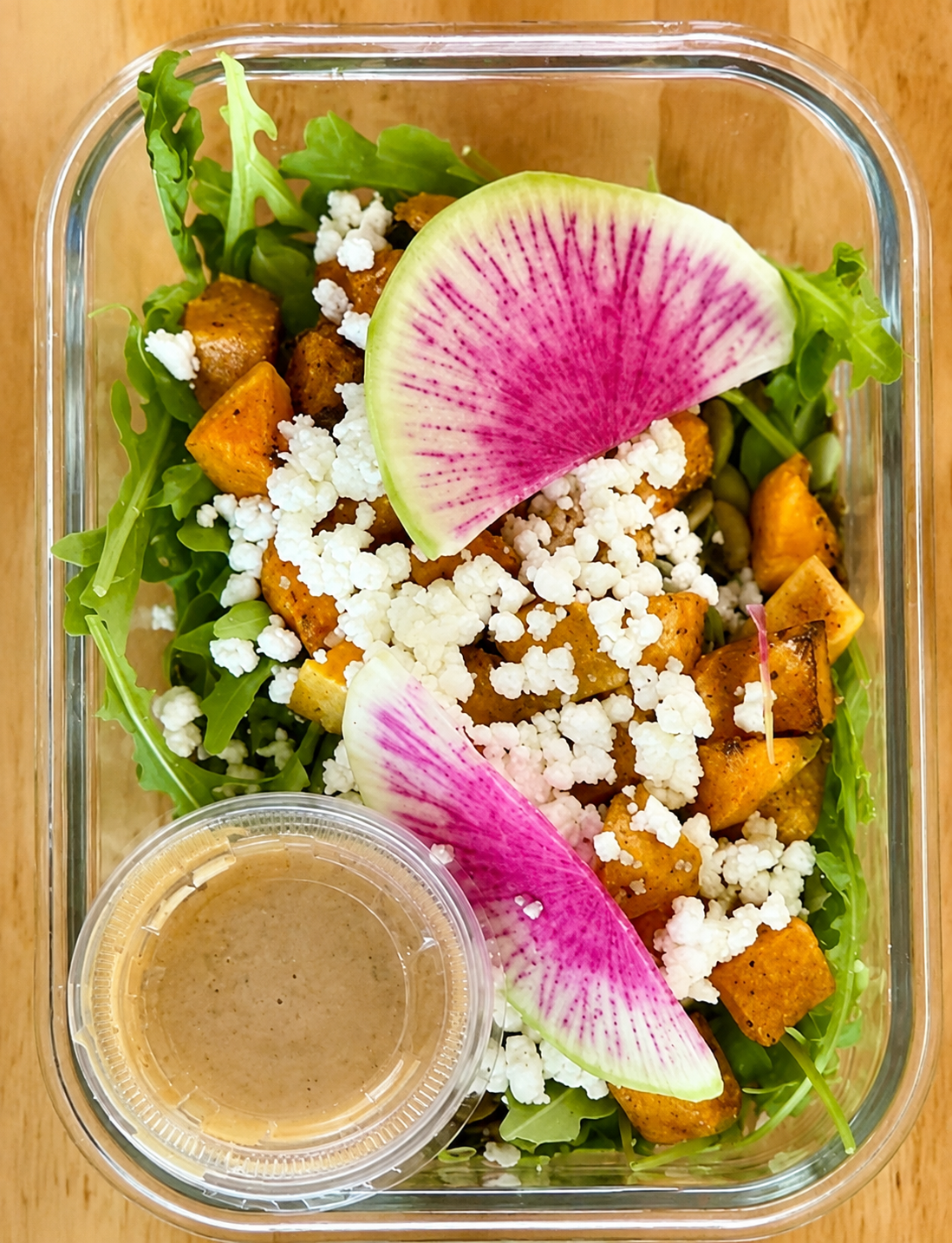 Salad with arugula, roasted sweet potatoes, crumbled cheese, two slices of watermelon radish, and a small container of salad dressing in a clear glass container.