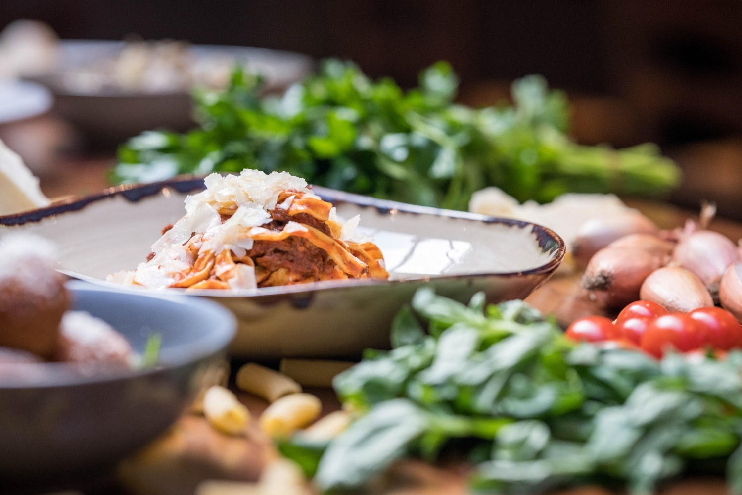 A close up spread of traditional Italian food, including pasta, basil and tomatoes, on a rustic wooden table.