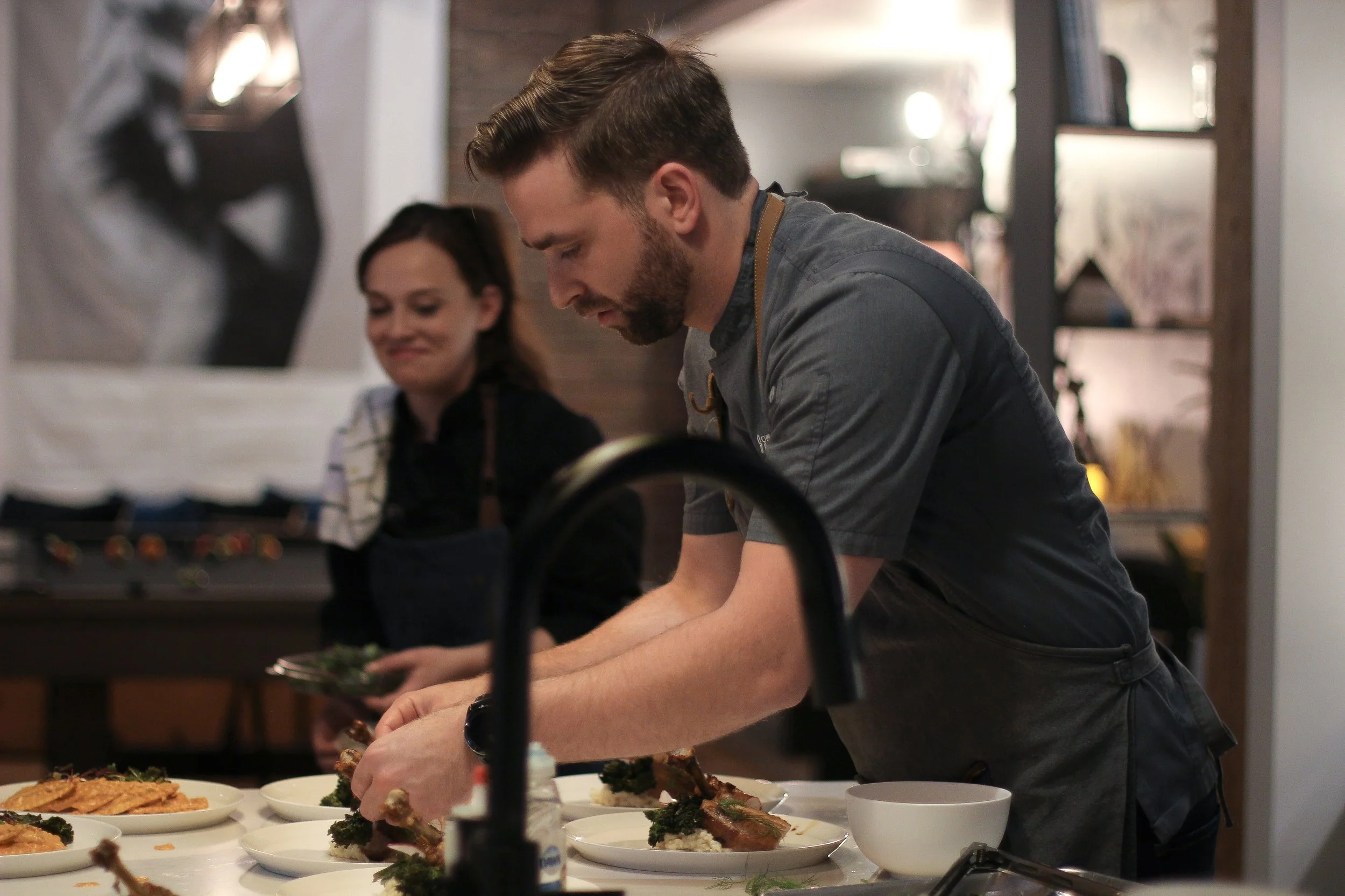 A man with a beard and short hair preparing food on a countertop in a kitchen, with a woman smiling in the background, surrounded by plates of food and kitchen utensils.