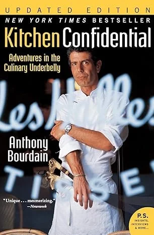 Book cover of 'Kitchen Confidential' by Anthony Bourdain, featuring a portrait of Bourdain in a white chef's coat with a dark background and neon lights.