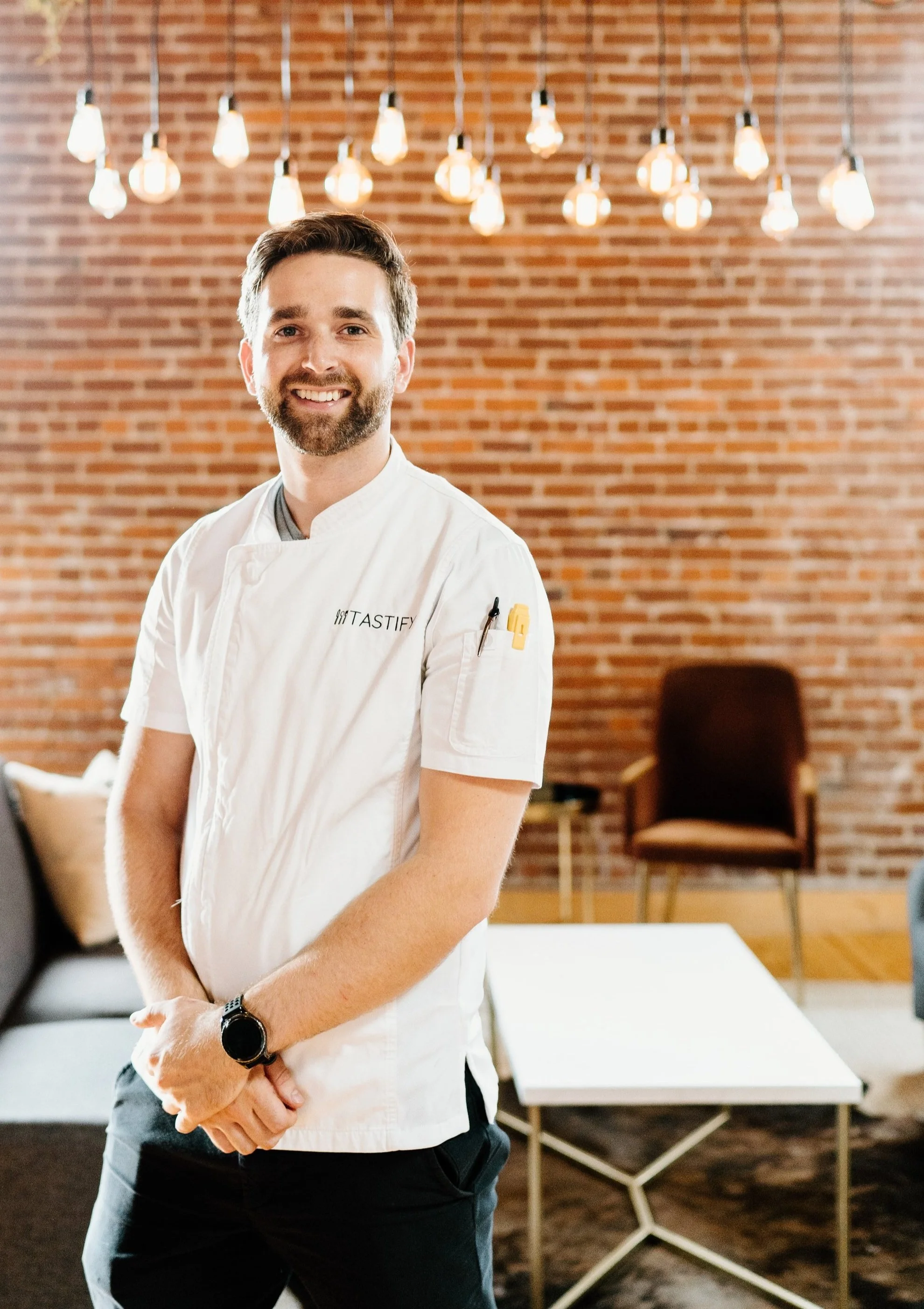 A smiling man in a white chef's coat standing in a room with a brick wall background and hanging light bulbs.