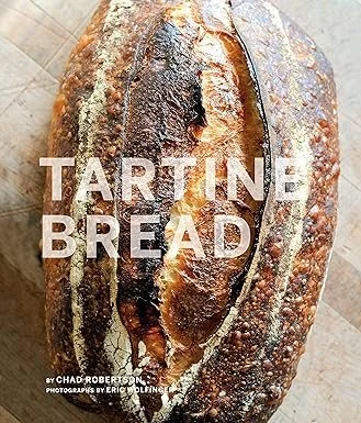 A loaf of tartine bread with a dark crust and scored top, placed on a wooden surface, with the words 'Tartine Bread' overlaid on the image.