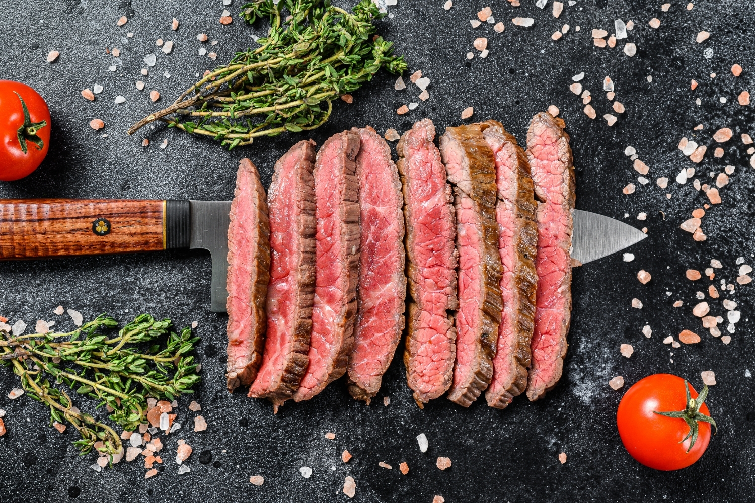 A perfectly cooked medium-rare steak sliced and sitting on a chefs knife over a slate chopping board with sea salt and fresh rosemary.