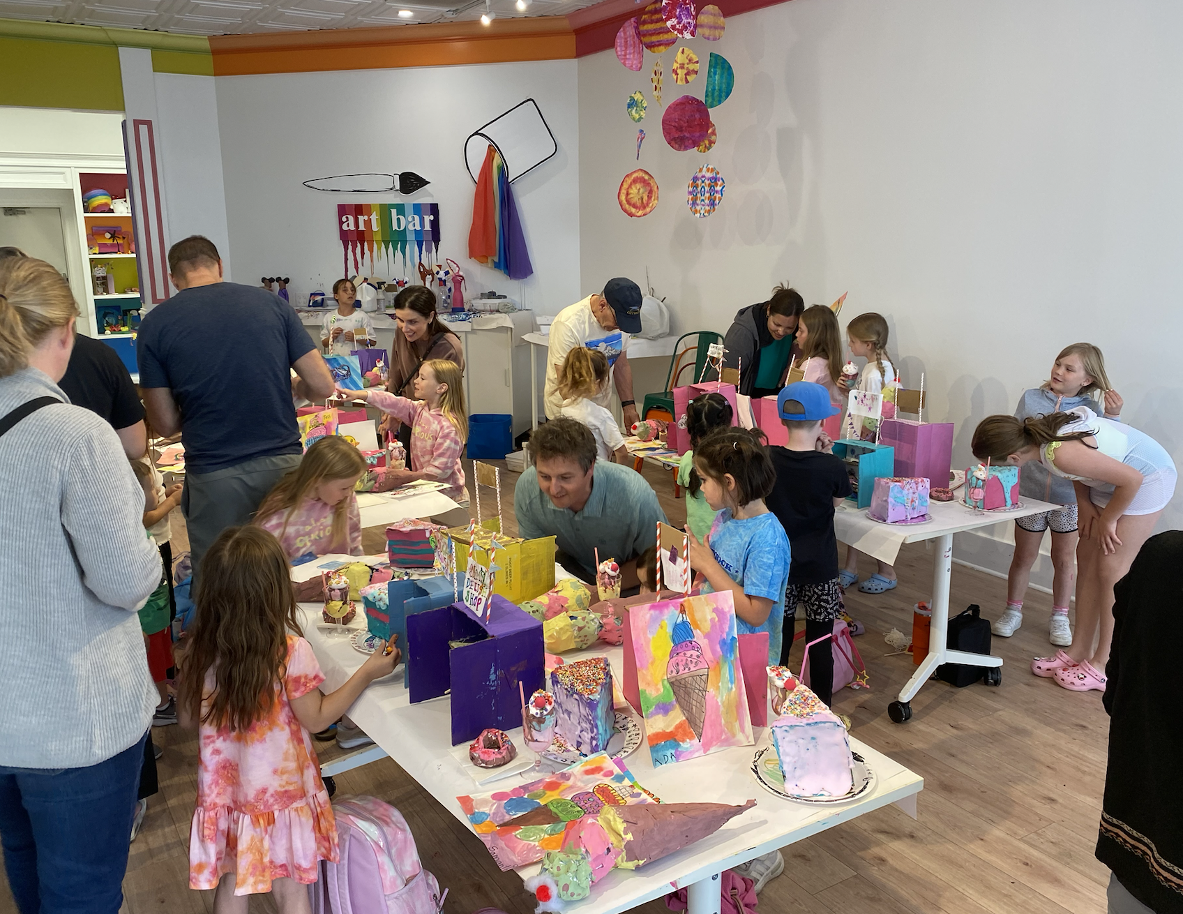 kids showing parents their summer art camp artwork they created at camp. art work is displayed on tables as an art show.