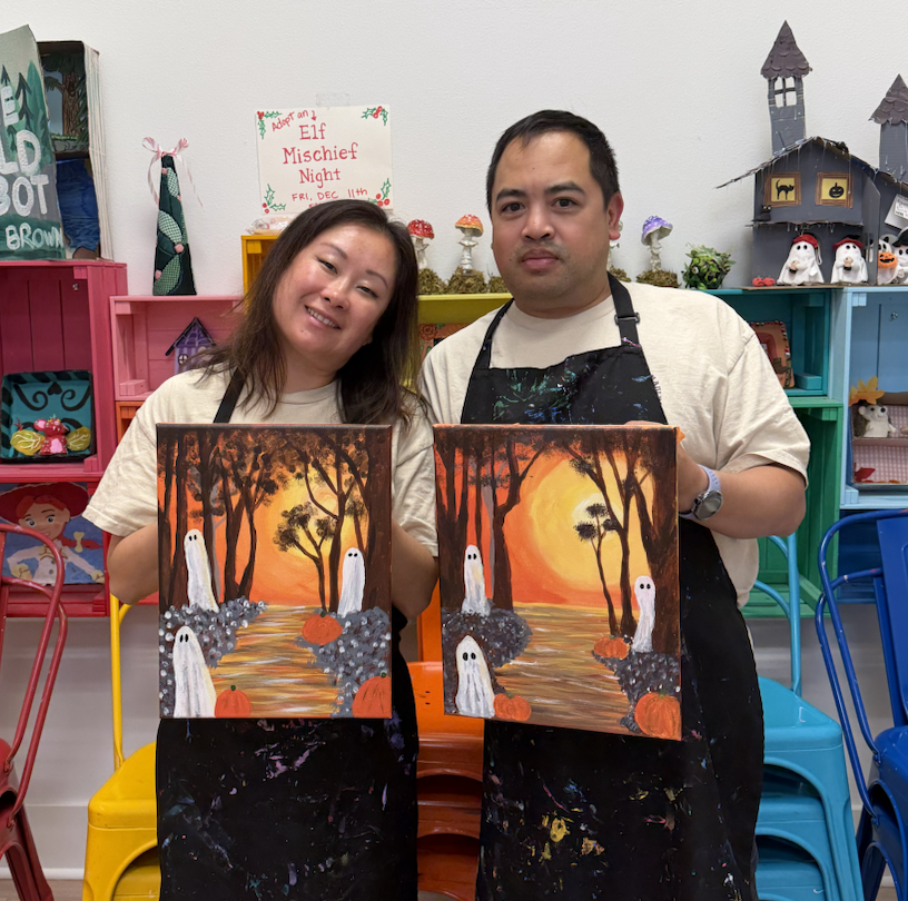 Two adults holding their finished Halloween paintings featuring ghosts, pumpkins, and an orange sunset at Whimsy Wees Art Studio.