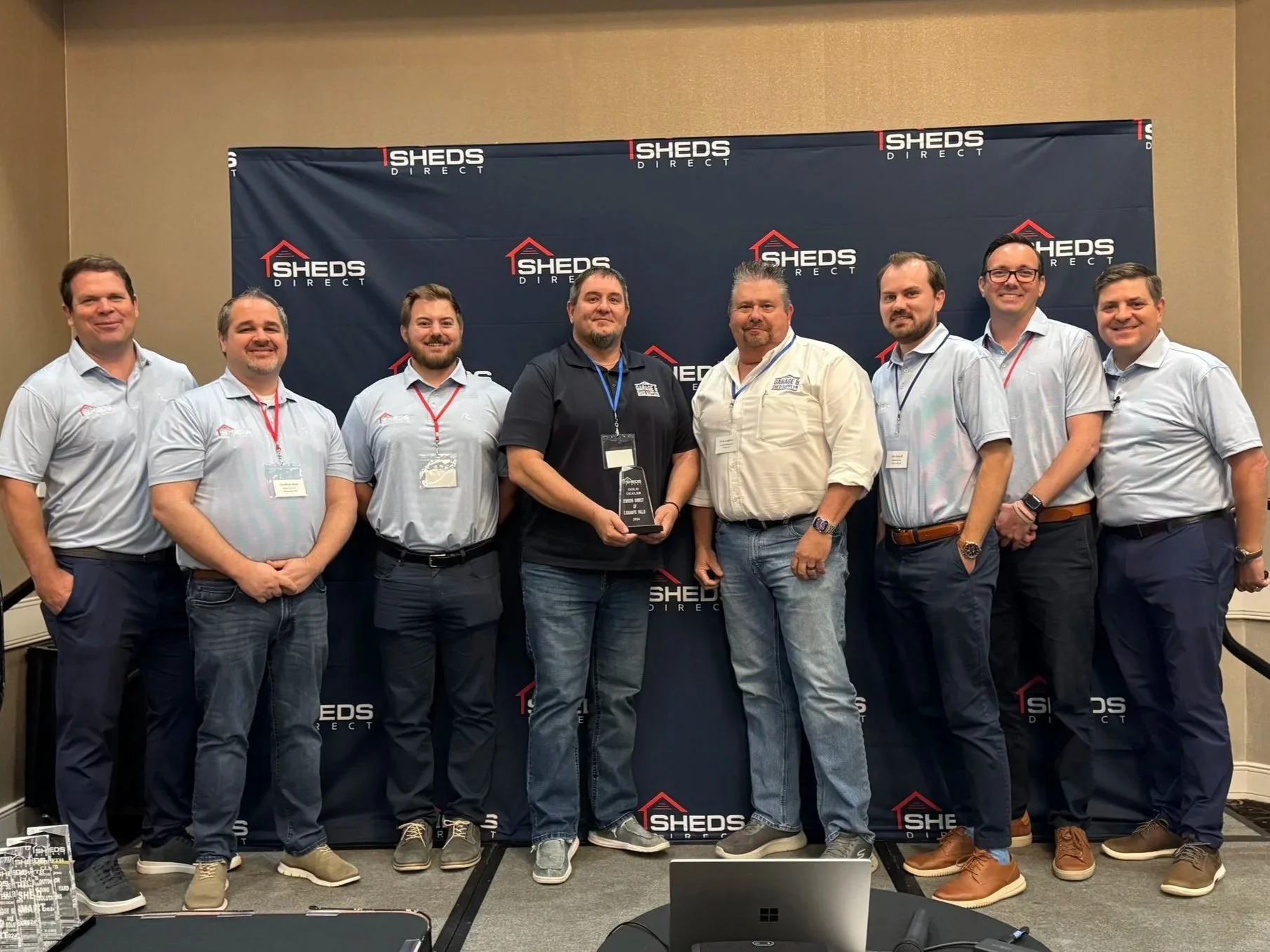 Group of nine men standing on a stage in front of a backdrop with the SHEDS Direct logo, at an awards event. One man is holding a trophy.