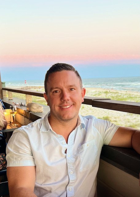 Man smiling at a seaside restaurant during sunset, sitting at a table with a view of the beach and ocean.