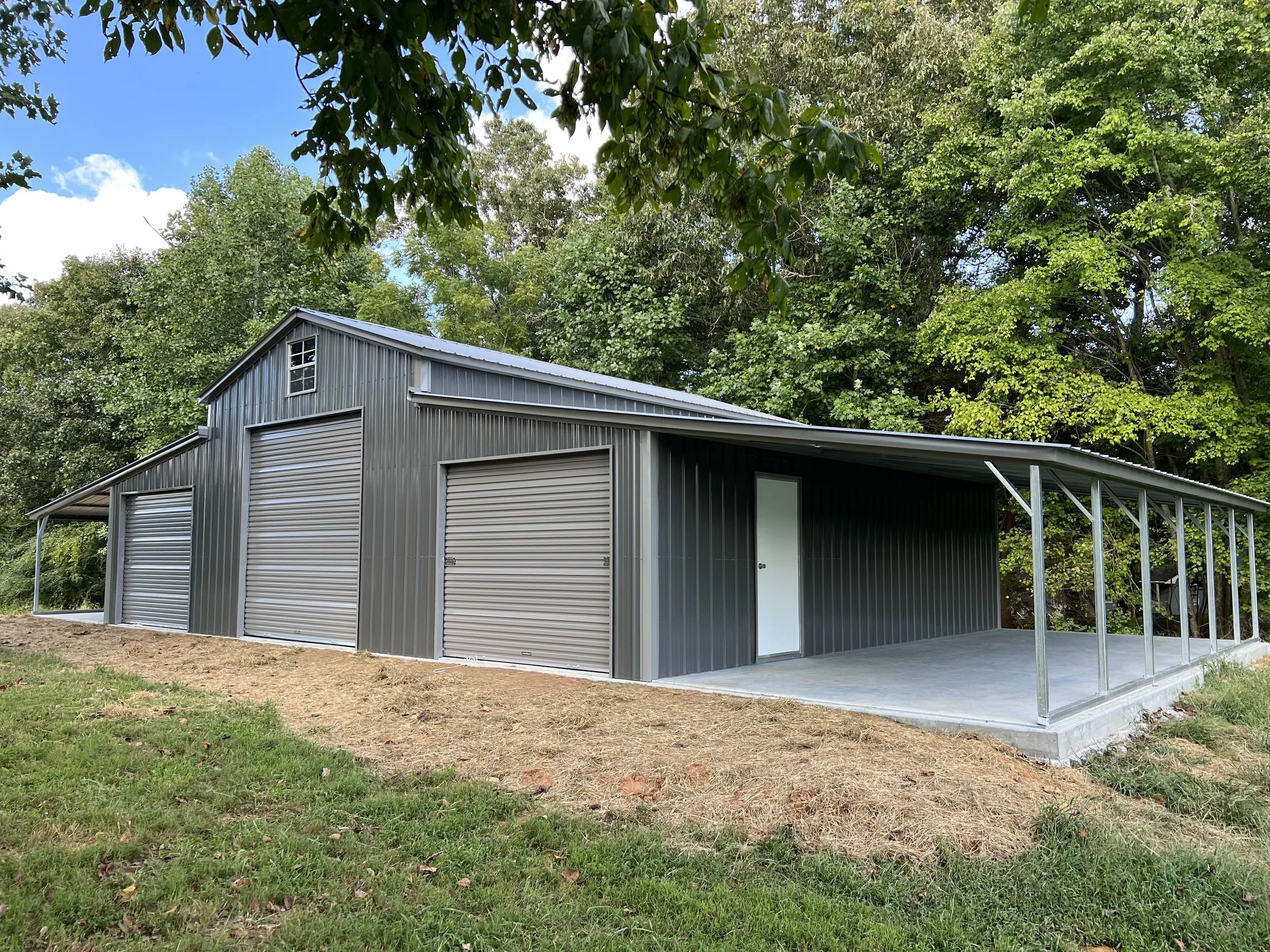 Dark gray barn with garage door openings, second story window, white door, and covered area; by Garage & Shed Supply.