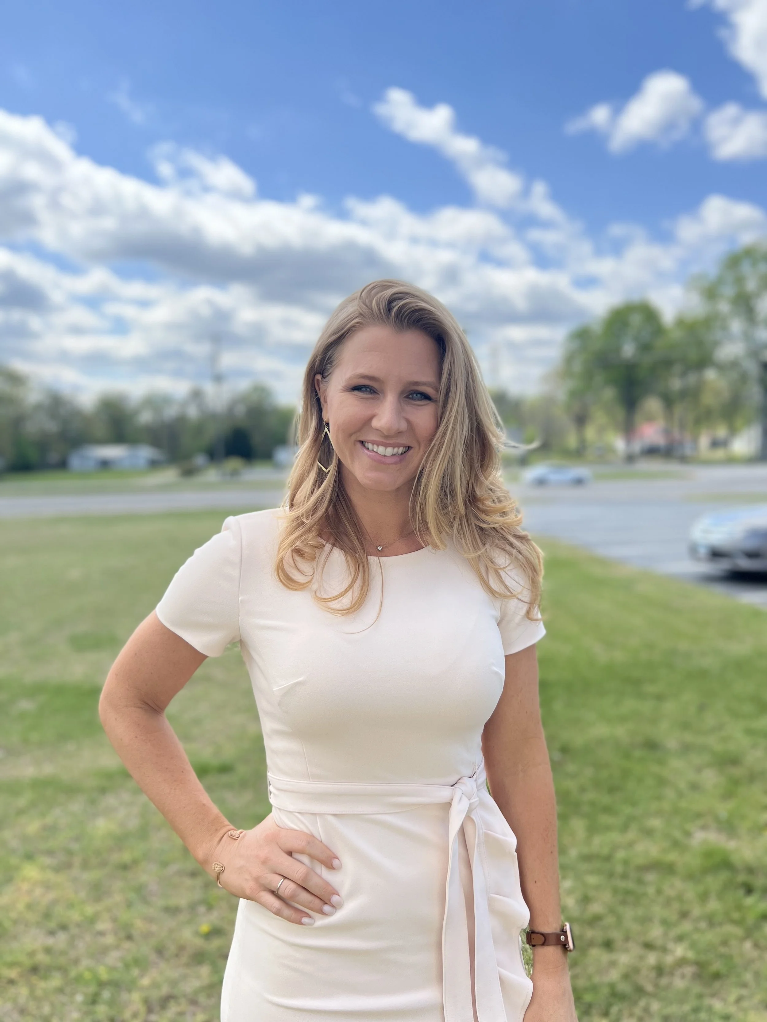 A smiling woman with blonde hair wearing a white dress standing outdoors on a grassy area on a partly cloudy day.