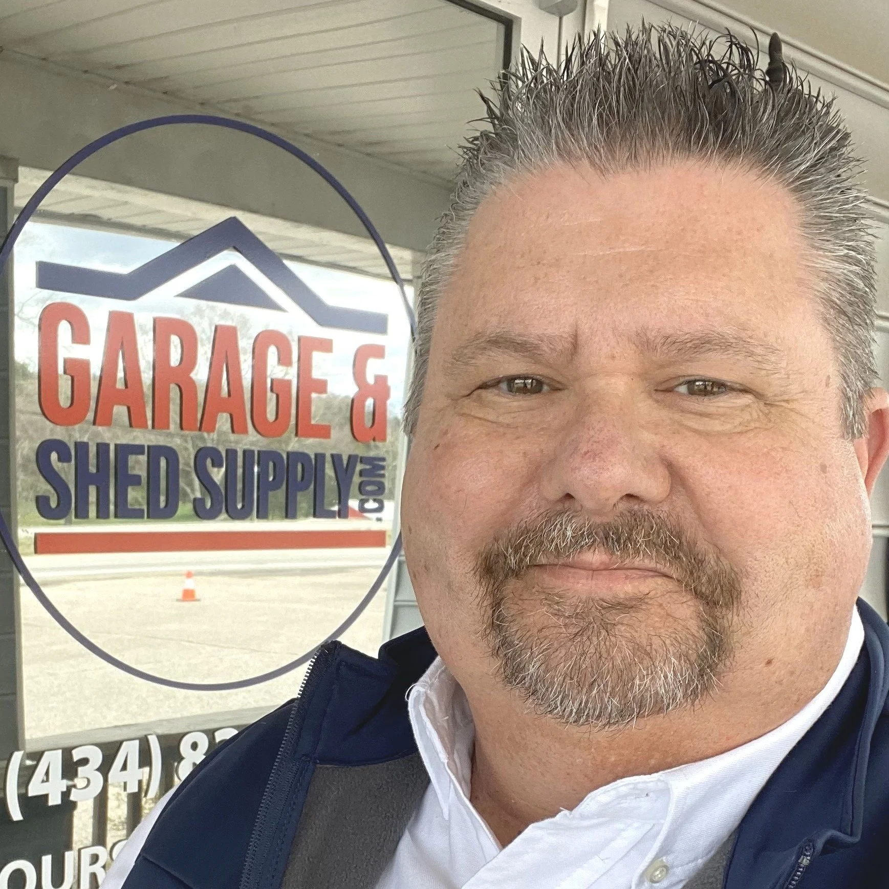 A man taking a selfie outside a store with a sign reading "Garage & Shed Supply" in the background.