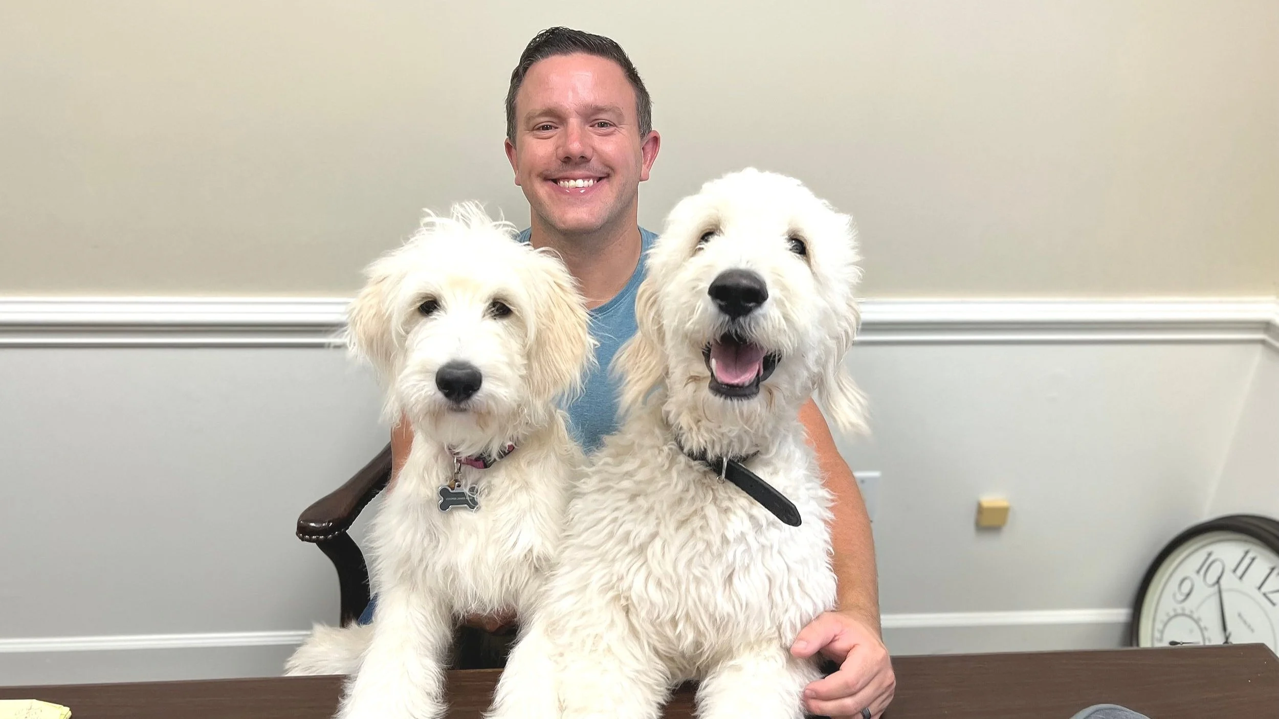 A man sitting at a table, smiling, with two fluffy white dogs in front of him. The man is wearing a blue shirt, and the dogs are sitting on the table.