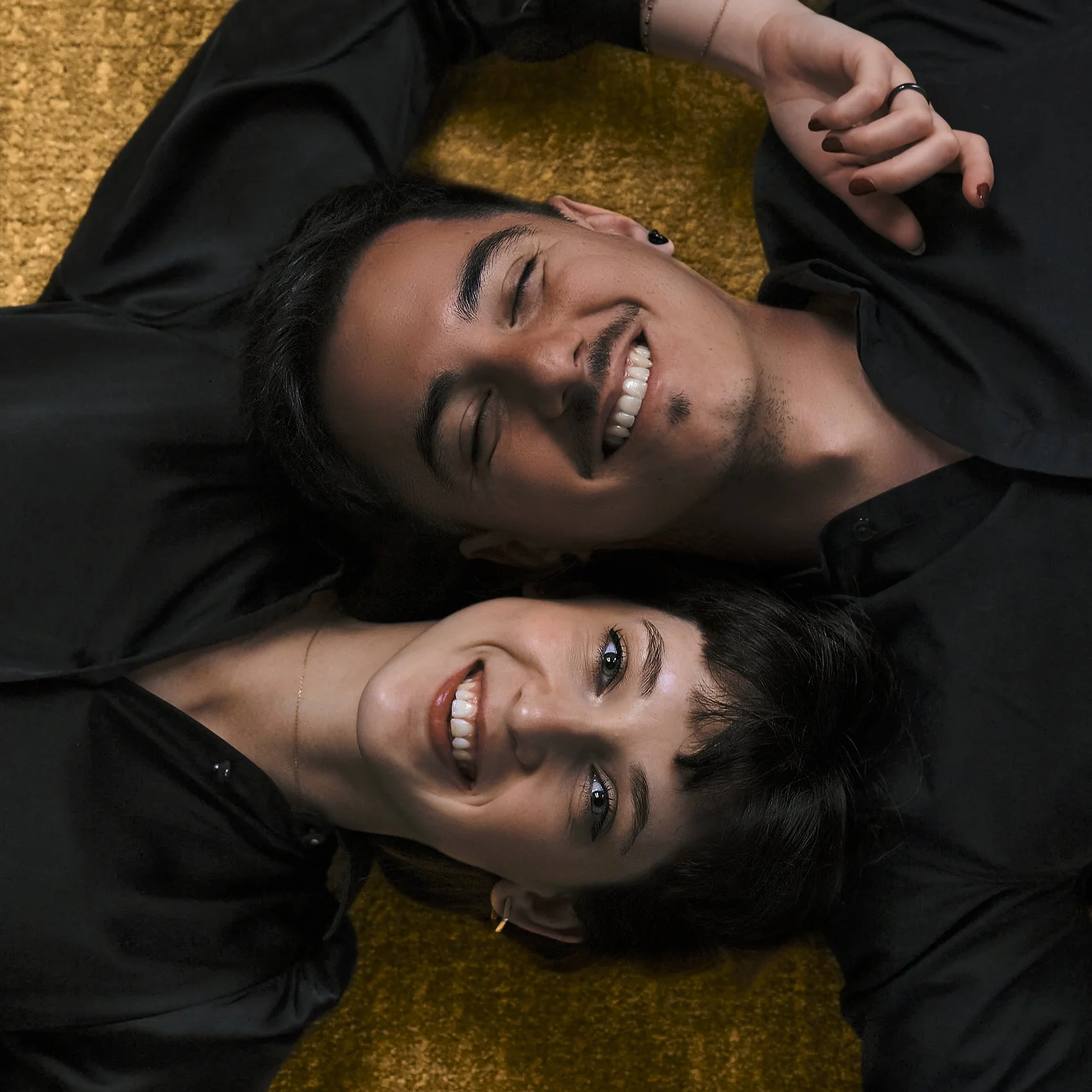 Deux personnes, un homme et une femme, allongés sur un tapis avec un fond jaune. Tous deux souriants et regardant en direction de l'appareil photo.