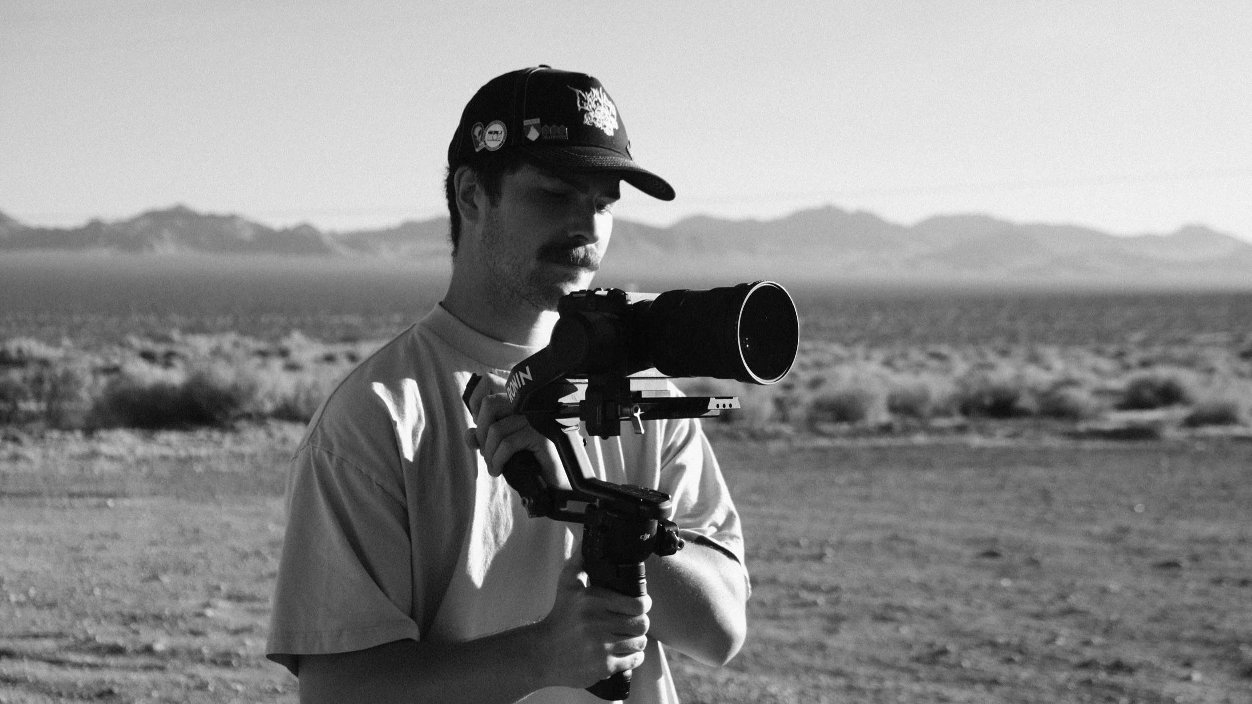 A man with a mustache wearing a baseball cap and a light-colored shirt is looking through a camera mounted on a tripod in a desert landscape with mountains in the background.