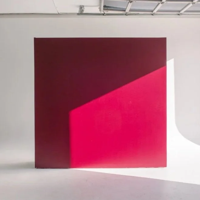 A large square canvas with a gradient from dark maroon at the top to bright pink at the bottom, leaning against a white wall. A shadow is cast on the canvas from an unseen light source.