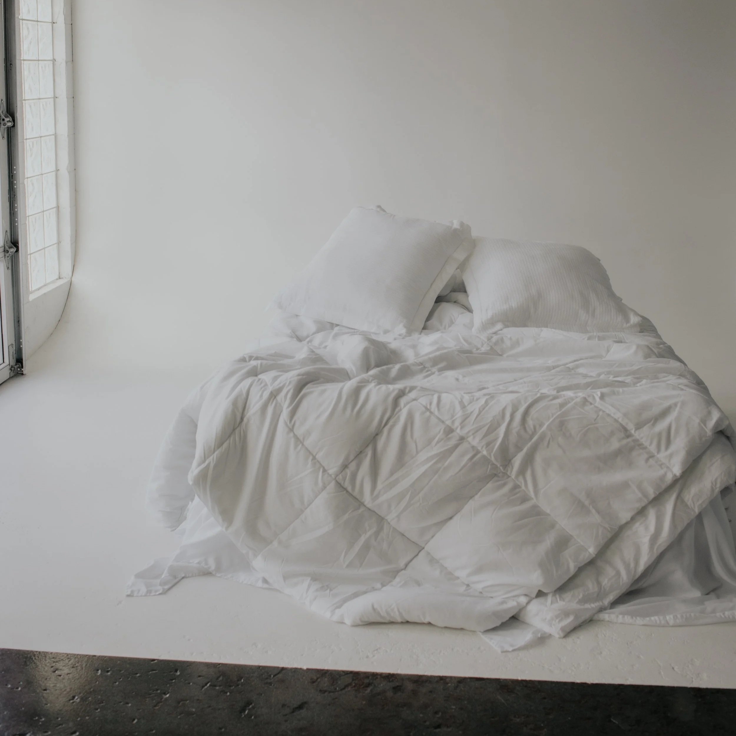 A neatly made bed with white linens and two pillows, positioned near a large window in a minimalistic room with white walls and a concrete floor.